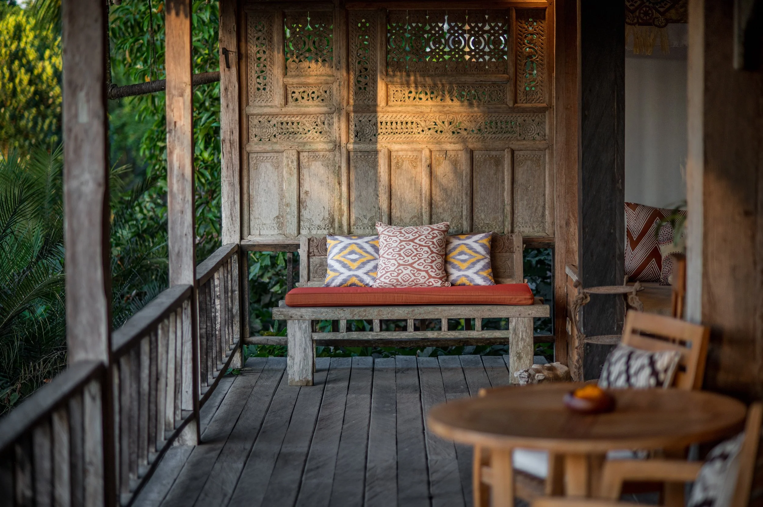 A rustic porch with a wooden bench and decorative pillows, surrounded by lush greenery and wooden architecture.