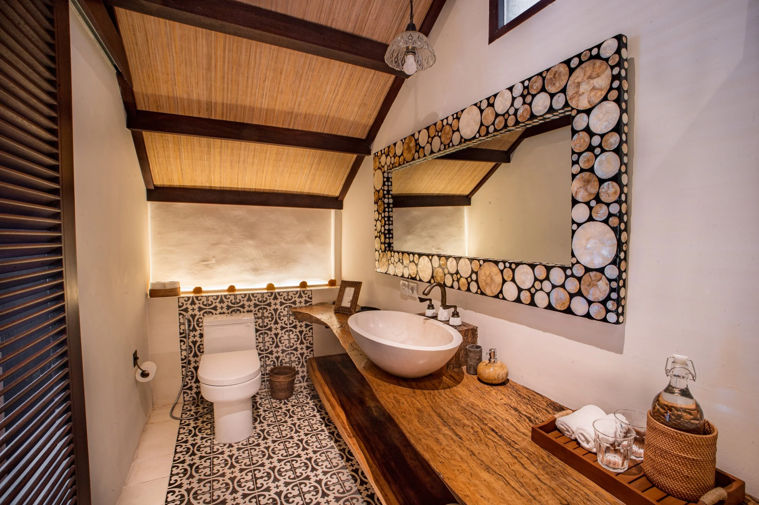 Bathroom with a rustic wooden countertop, a vessel sink, patterned floor and wall tiles, a toilet, a mirror with a log frame, and a decorative ceiling with exposed wood beams.