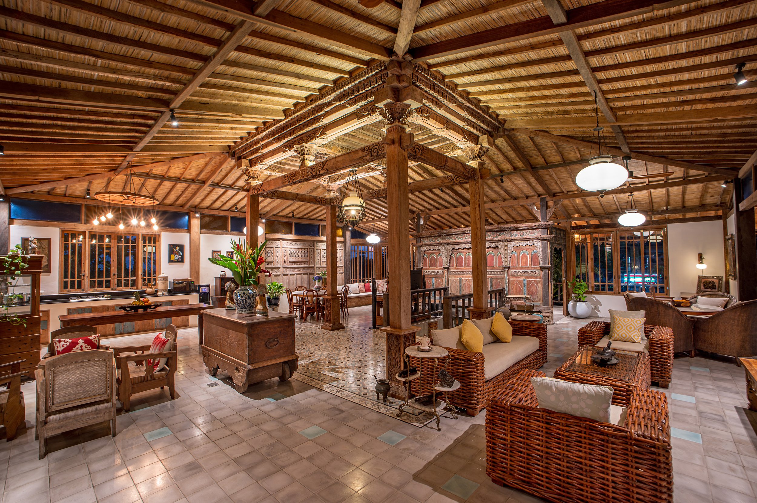 Interior of a spacious, well-lit living room with a wooden ceiling, multiple pendant lights, and a mix of modern and rustic furniture, including wicker chairs, a sofa with yellow pillows, and decorative plants.