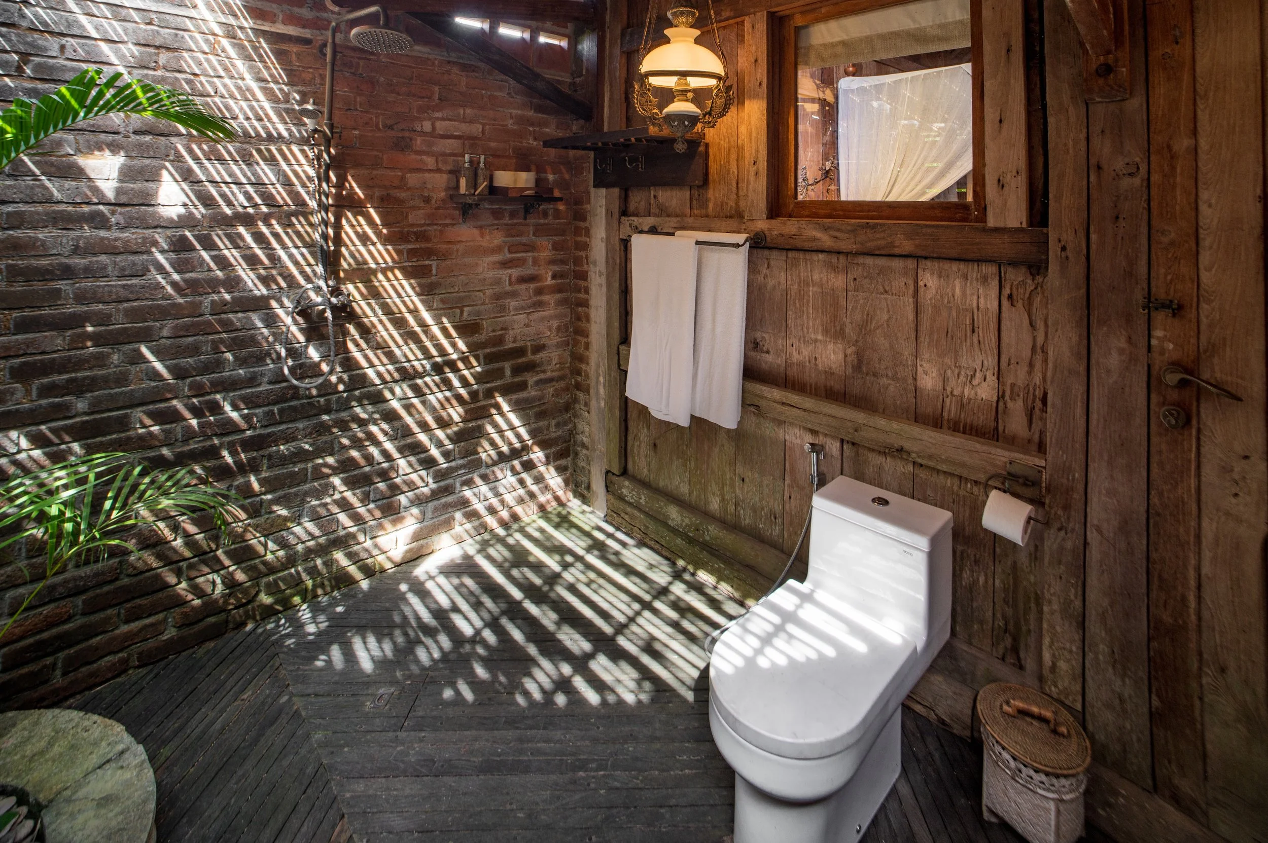 An outdoor bathroom with rustic wooden walls, a toilet, a small wicker basket, a window with a curtain, a towel rack with a white towel, and a showerhead mounted on a brick wall with sunlight casting patterns.