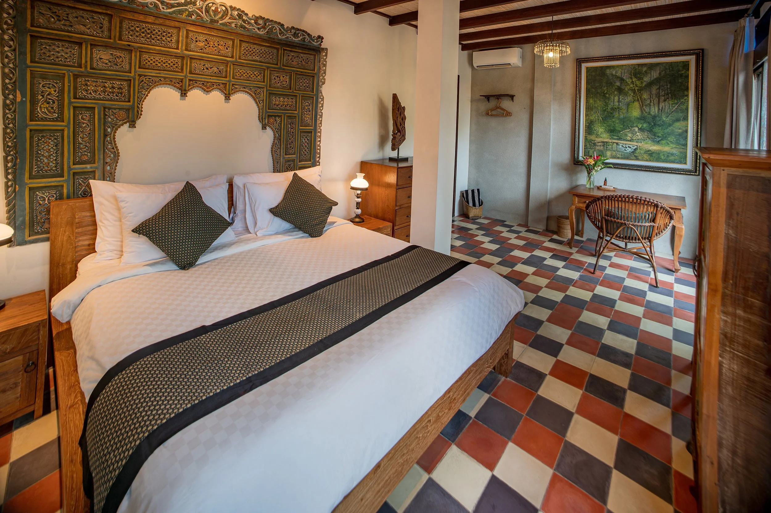 Hotel room with a bed, decorative headboard, nightstand with lamp, wooden furniture, framed landscape painting, and checkered tile floor.