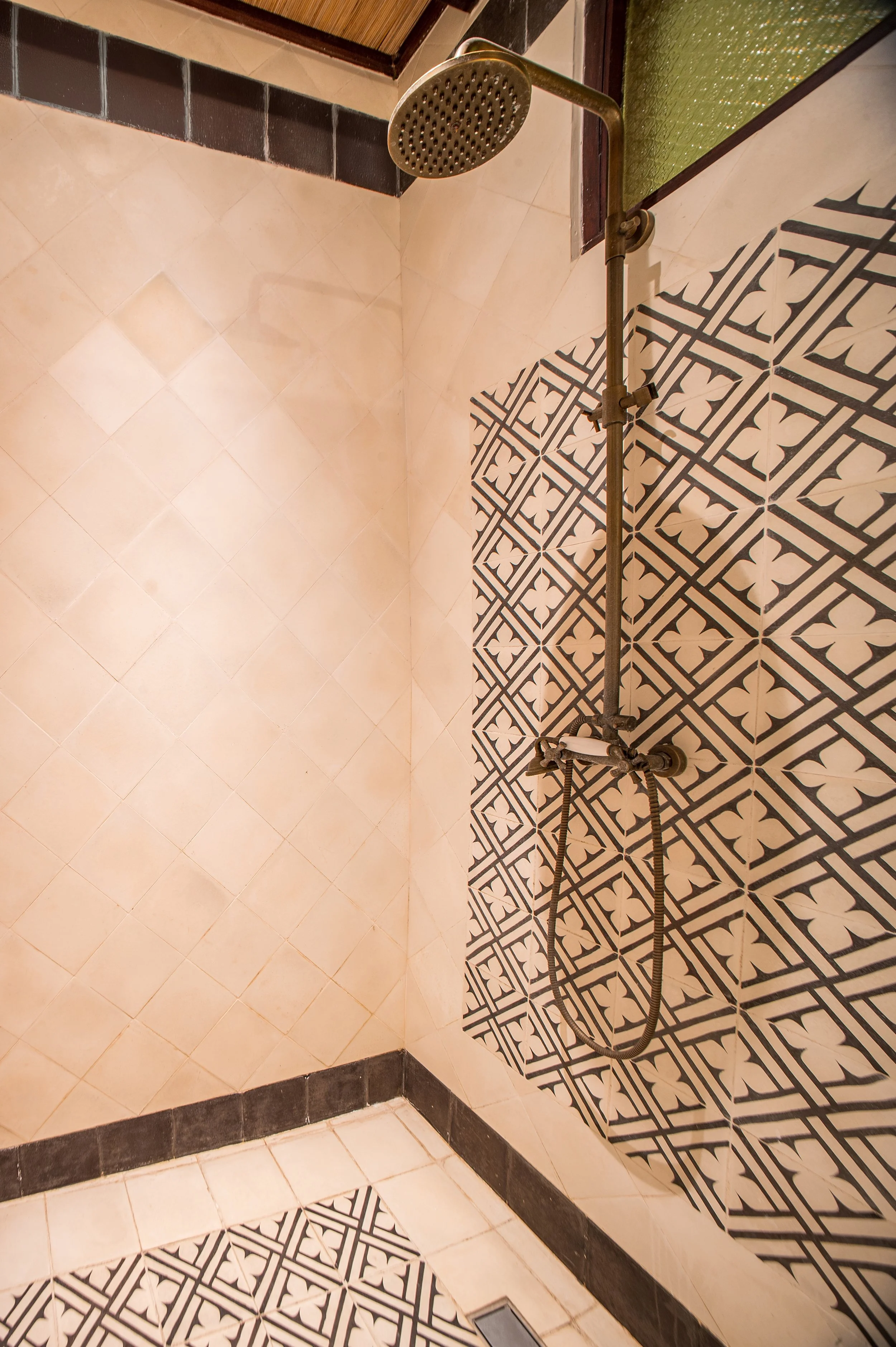 A shower area with patterned black and white floor tiles and plain beige wall tiles, featuring a wall-mounted shower head and black border tiles at the bottom.