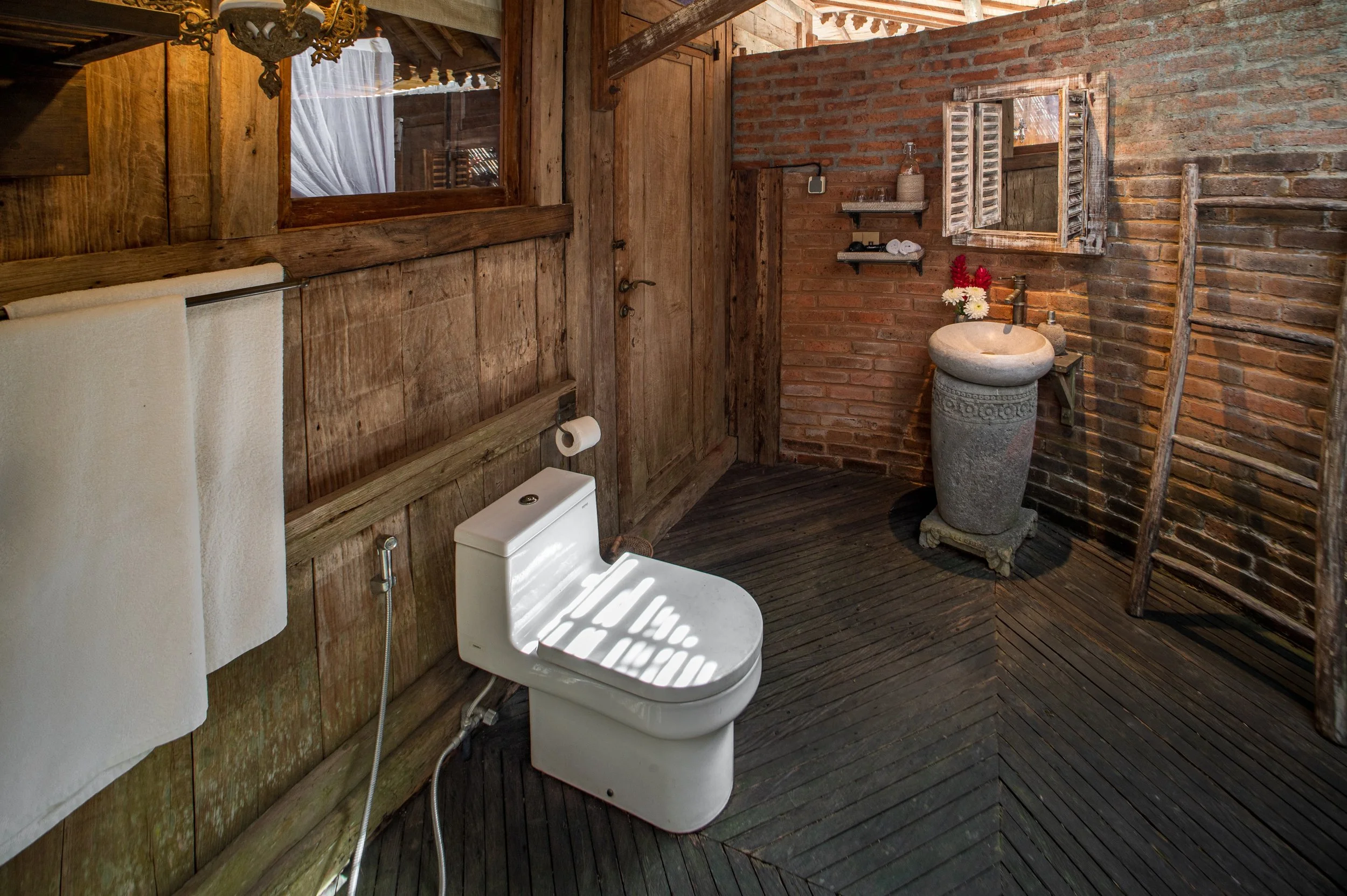 A rustic bathroom with brick and wooden walls, a stone sink with flowers, a wooden ladder, mirror with shutters, and a white toilet with a towel rack.