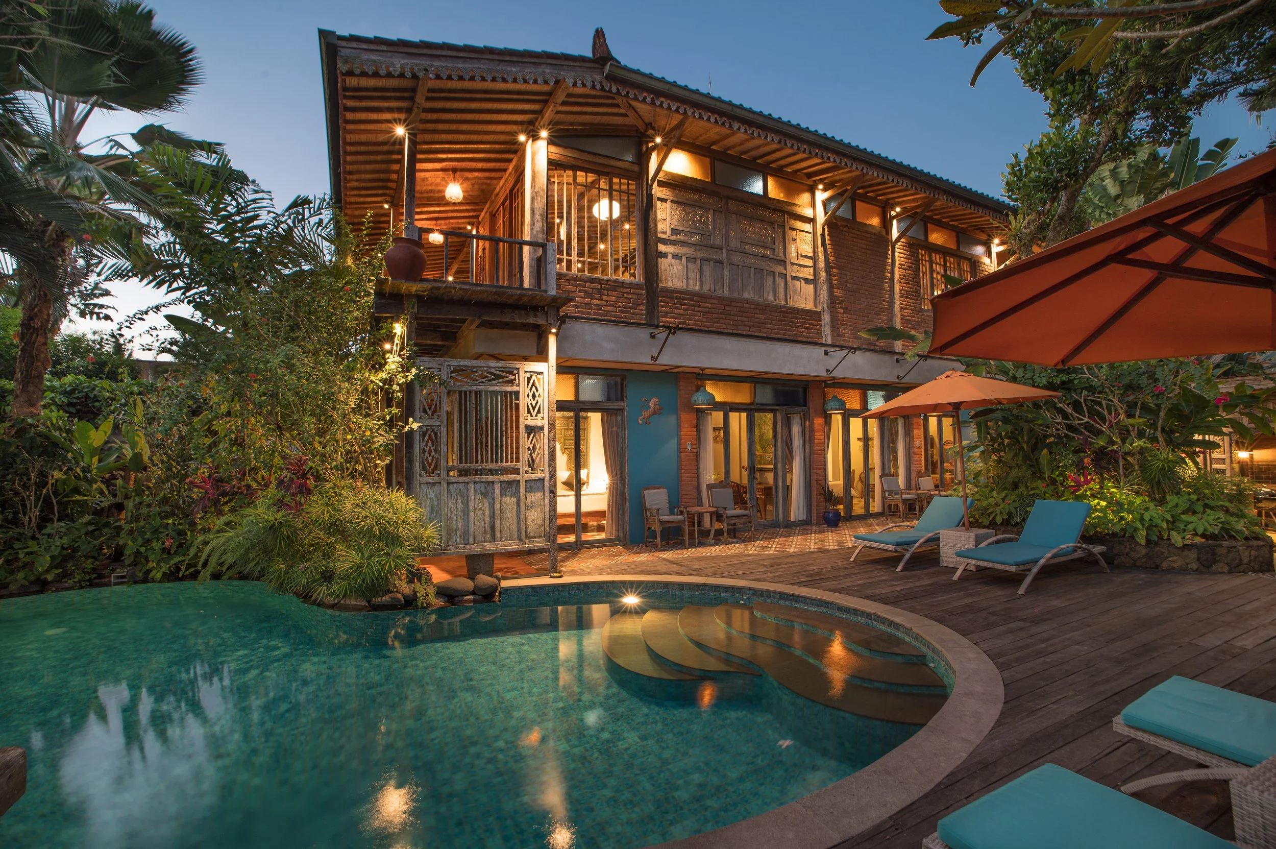 Exterior view of a two-story house with a pool and patio area, surrounded by lush greenery and trees, during twilight.