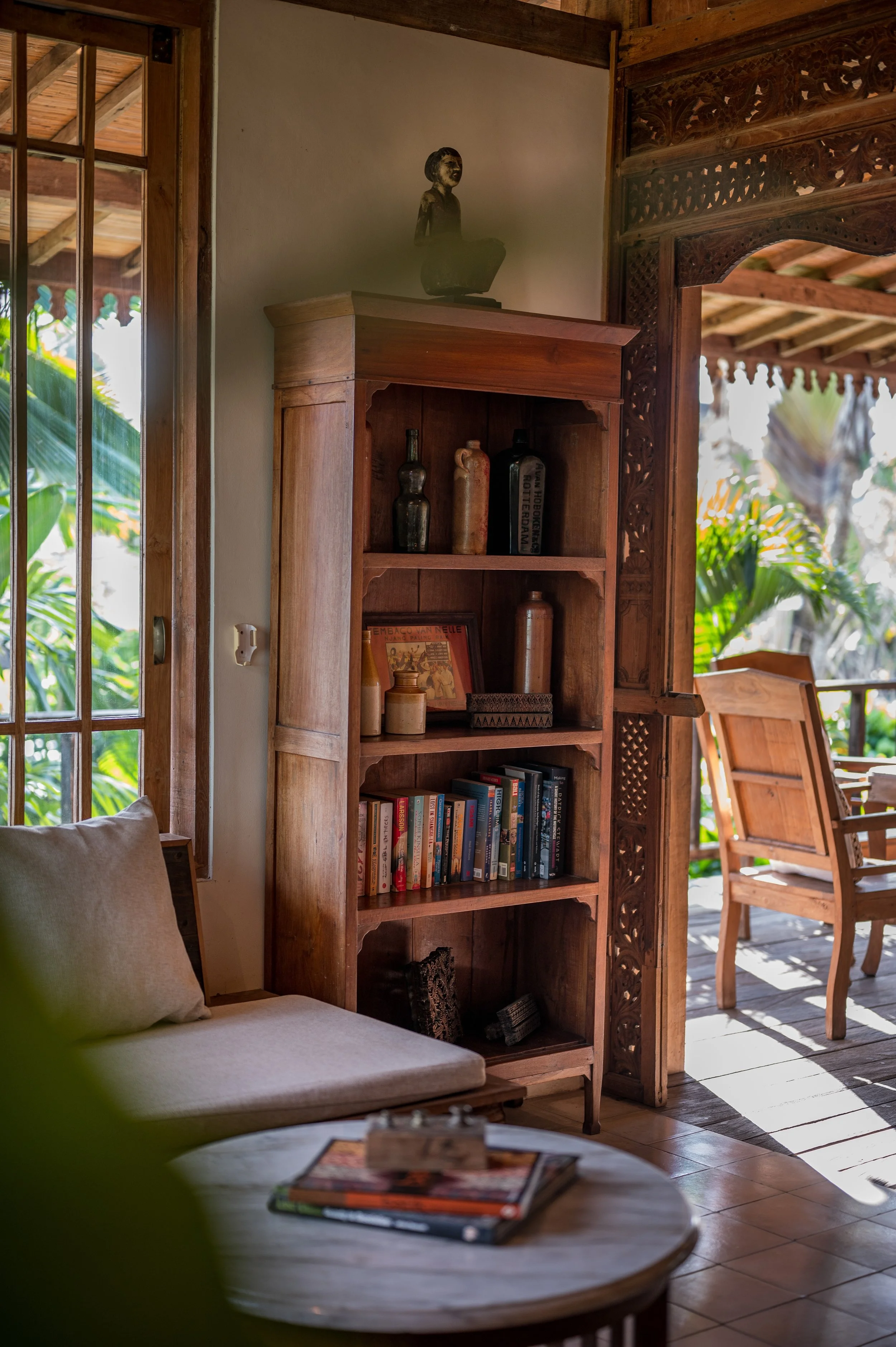 A cozy wooden interior with a white couch, a round coffee table with magazines, a wooden bookshelf with decorative items and books, a carved wooden partition, and a balcony with outdoor chairs and lush greenery.