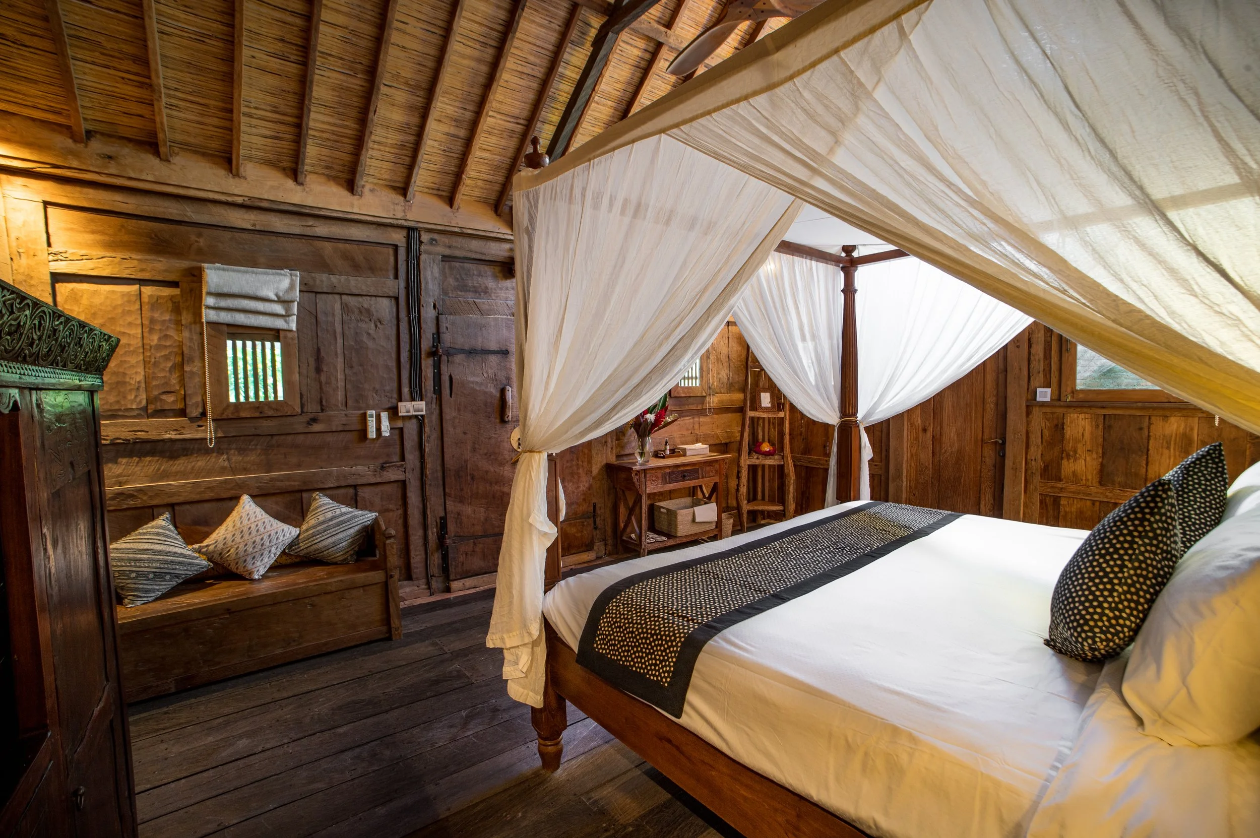 Interior of a rustic bedroom with a wooden four-poster bed, white canopy curtains, and a wooden wall with small windows.