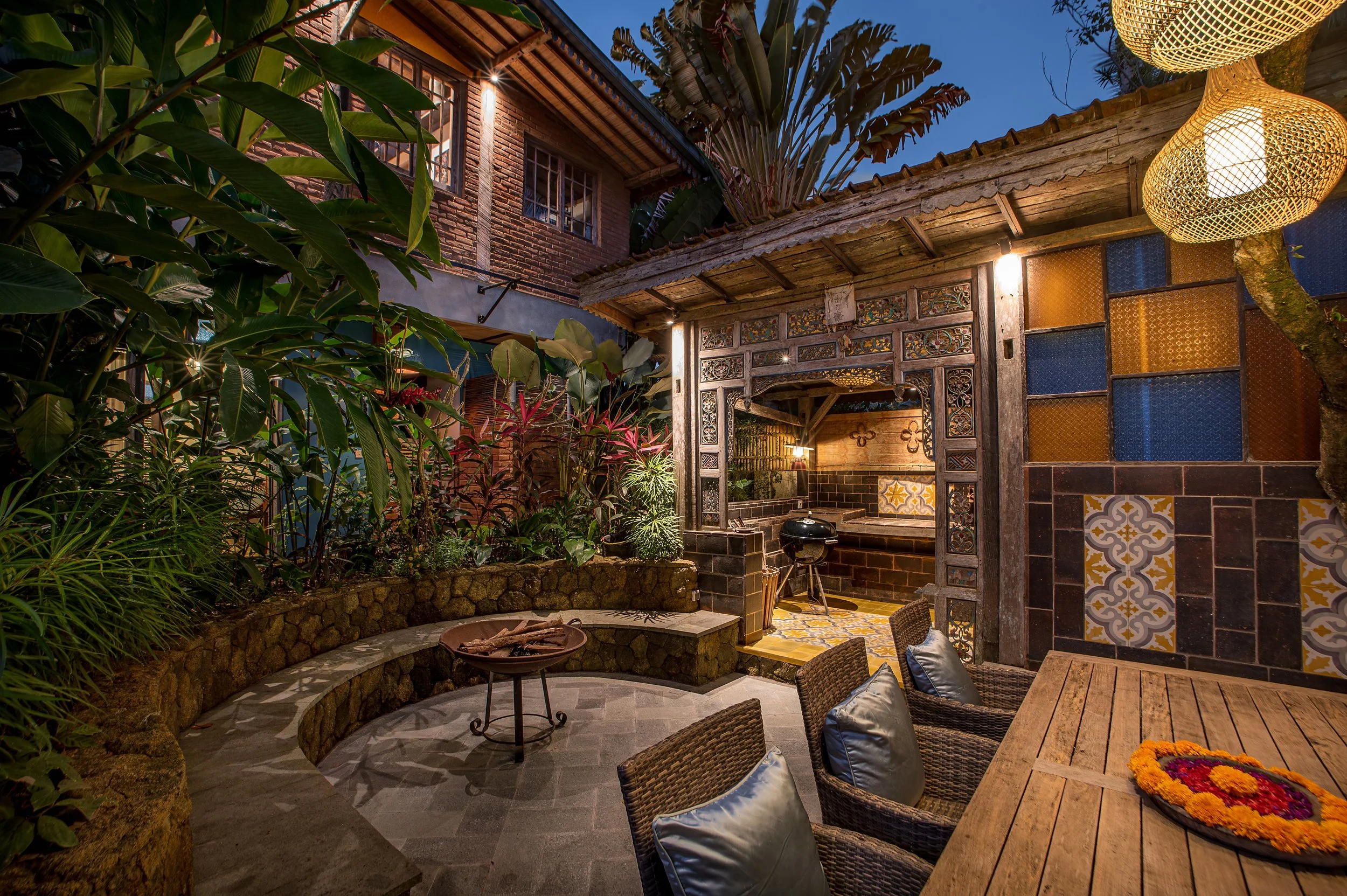 An outdoor patio area in the evening with string lights, lush green plants, a brick and wooden house, decorative tile accents, seating with chairs and cushions, a wooden table, and a fire pit.