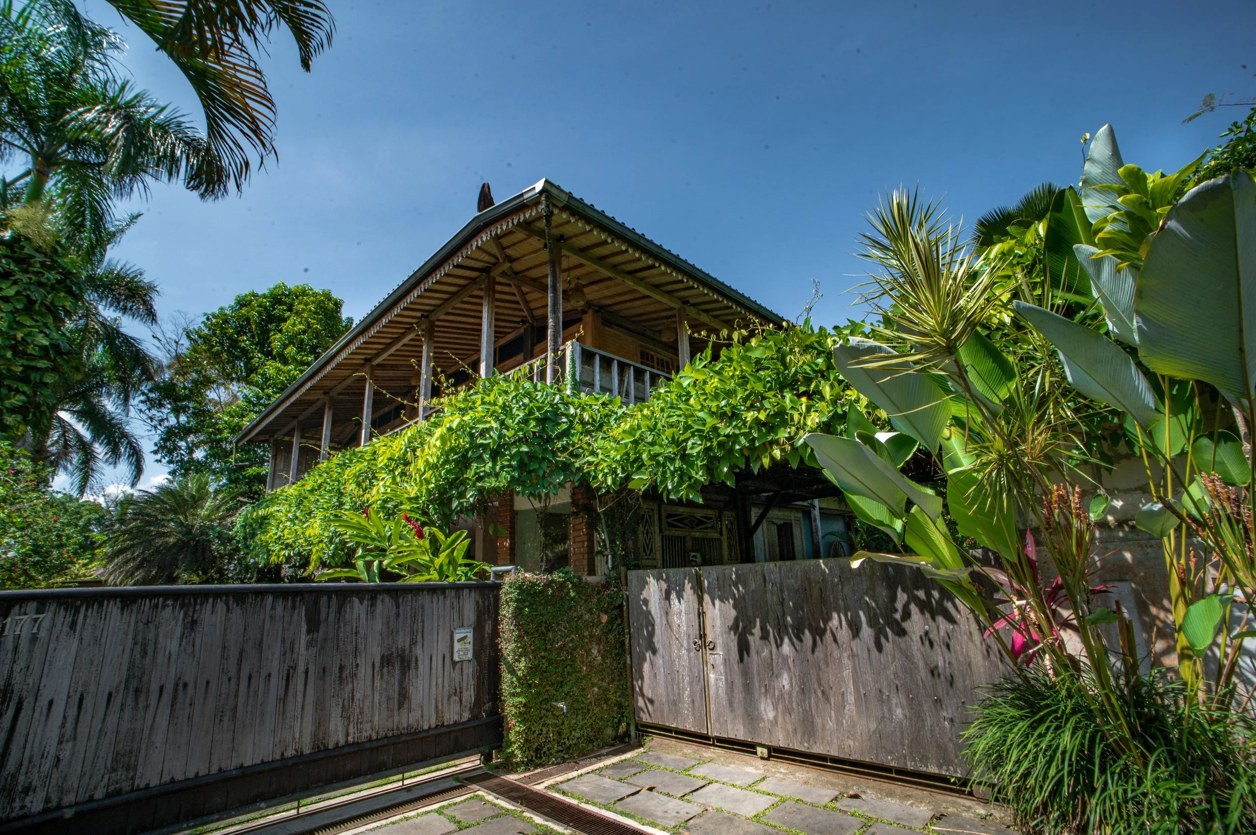 A two-story house atop a lush, green hillside, surrounded by dense tropical foliage, with a wooden fence and a gate in the foreground under a clear blue sky.