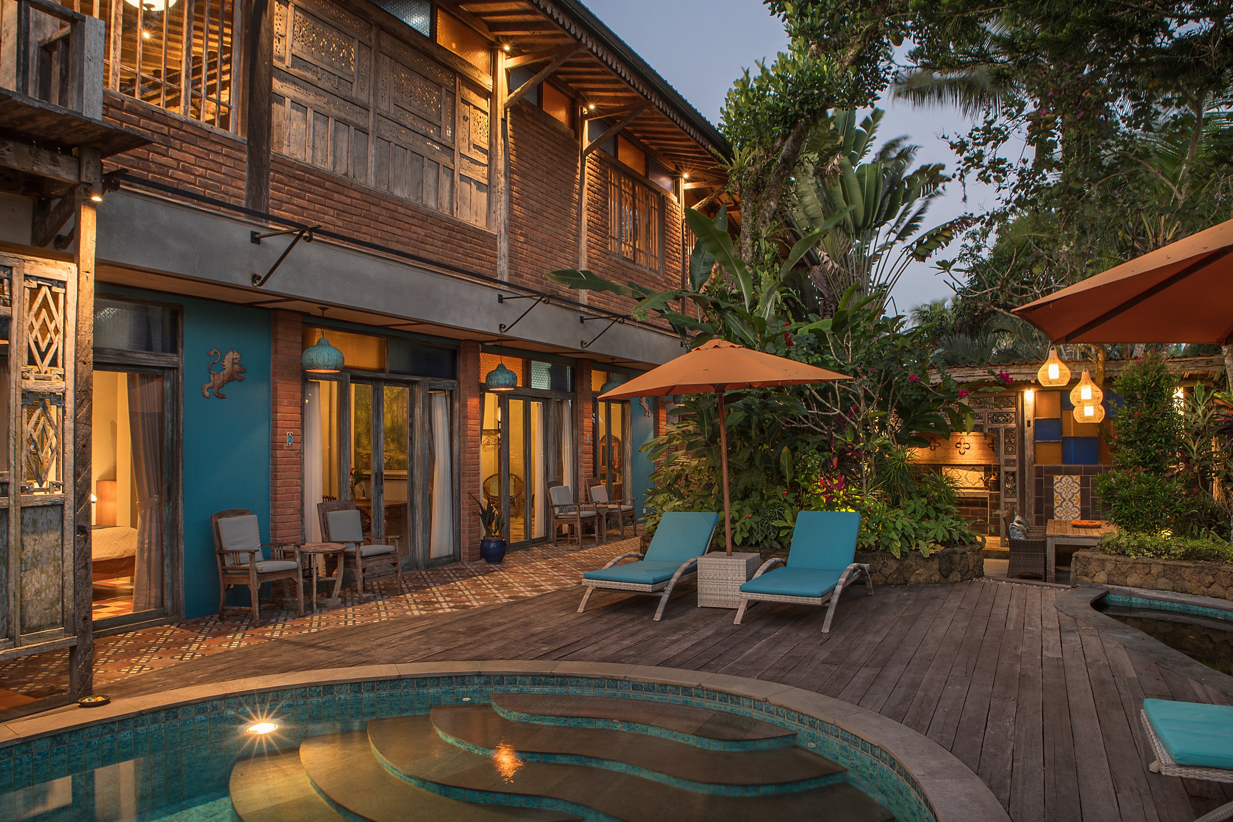 A backyard patio area featuring a swimming pool, lounge chairs with blue cushions, and an outdoor dining set under umbrellas. There are plants and trees surrounding the area, and a two-story house with wooden and brick accents, lit by outdoor lights.