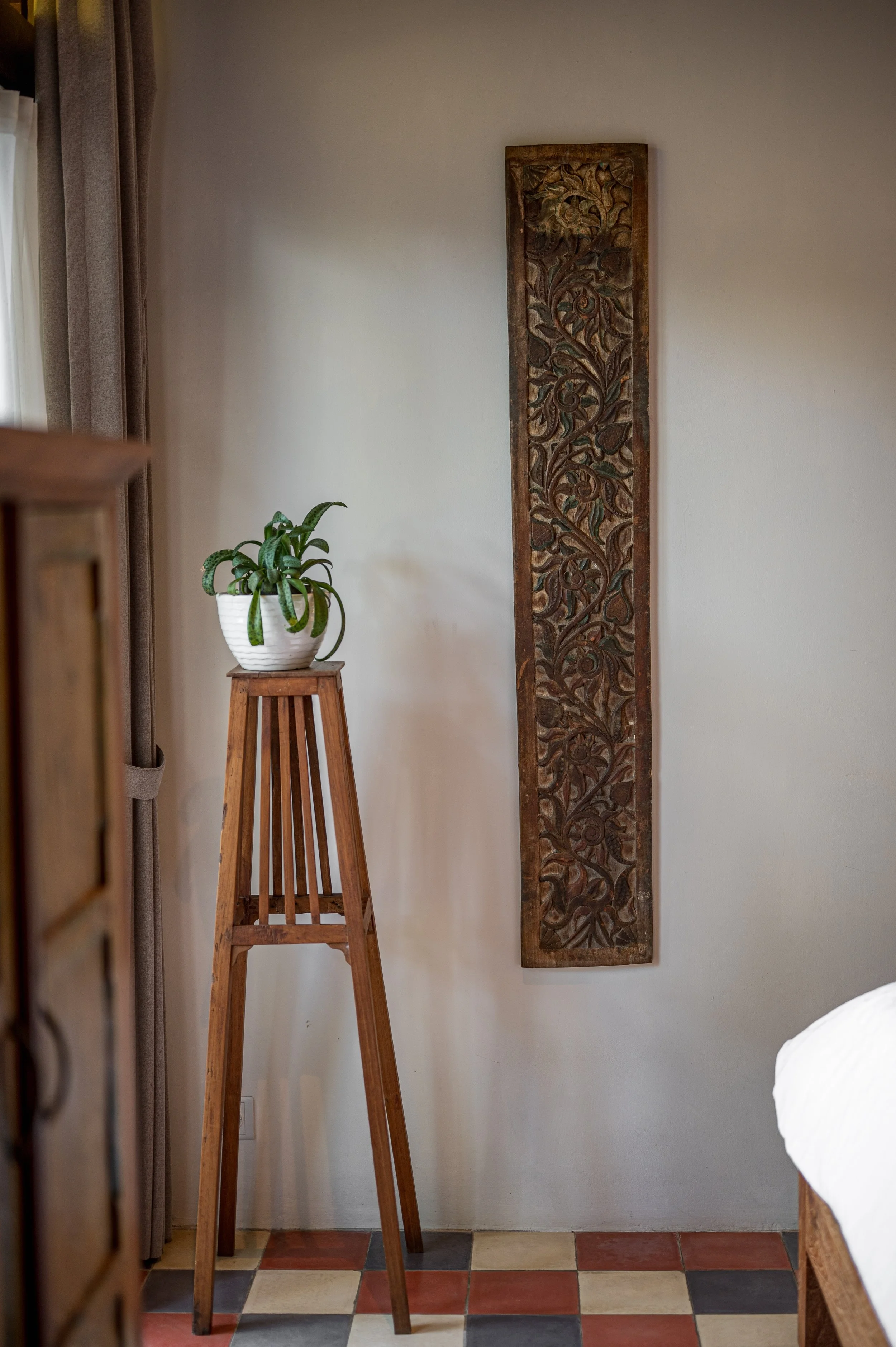 Interior of a room with a wooden plant stand holding a potted succulent plant, a decorative carved wooden panel on the wall, and a partially visible bed with a white duvet. There are beige curtains on the left and a floor with multicolored tiles.