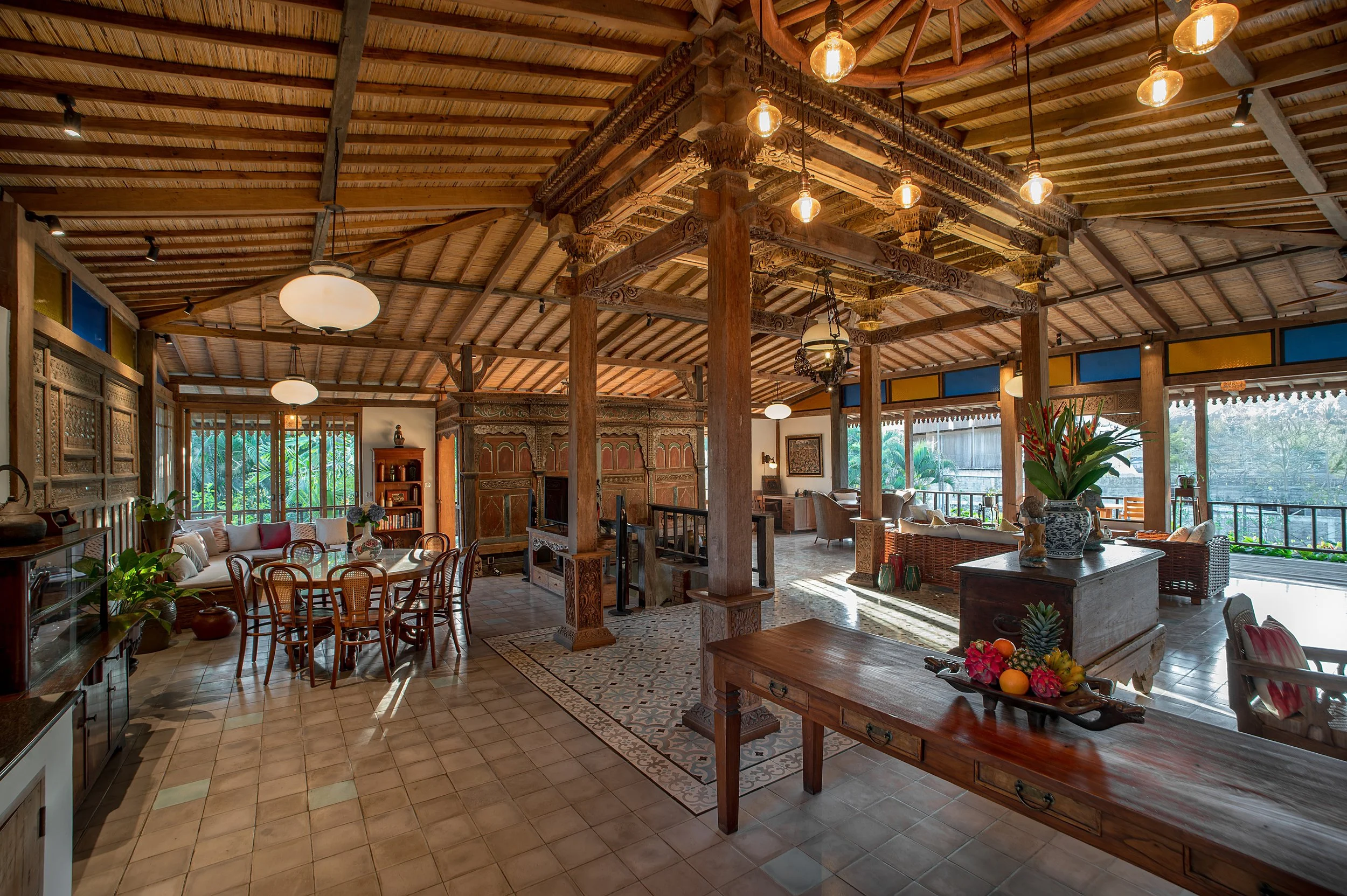 Spacious dining and living area with wooden furniture, decorative accents, and large windows showing greenery outside, under a wooden ceiling with hanging lights.
