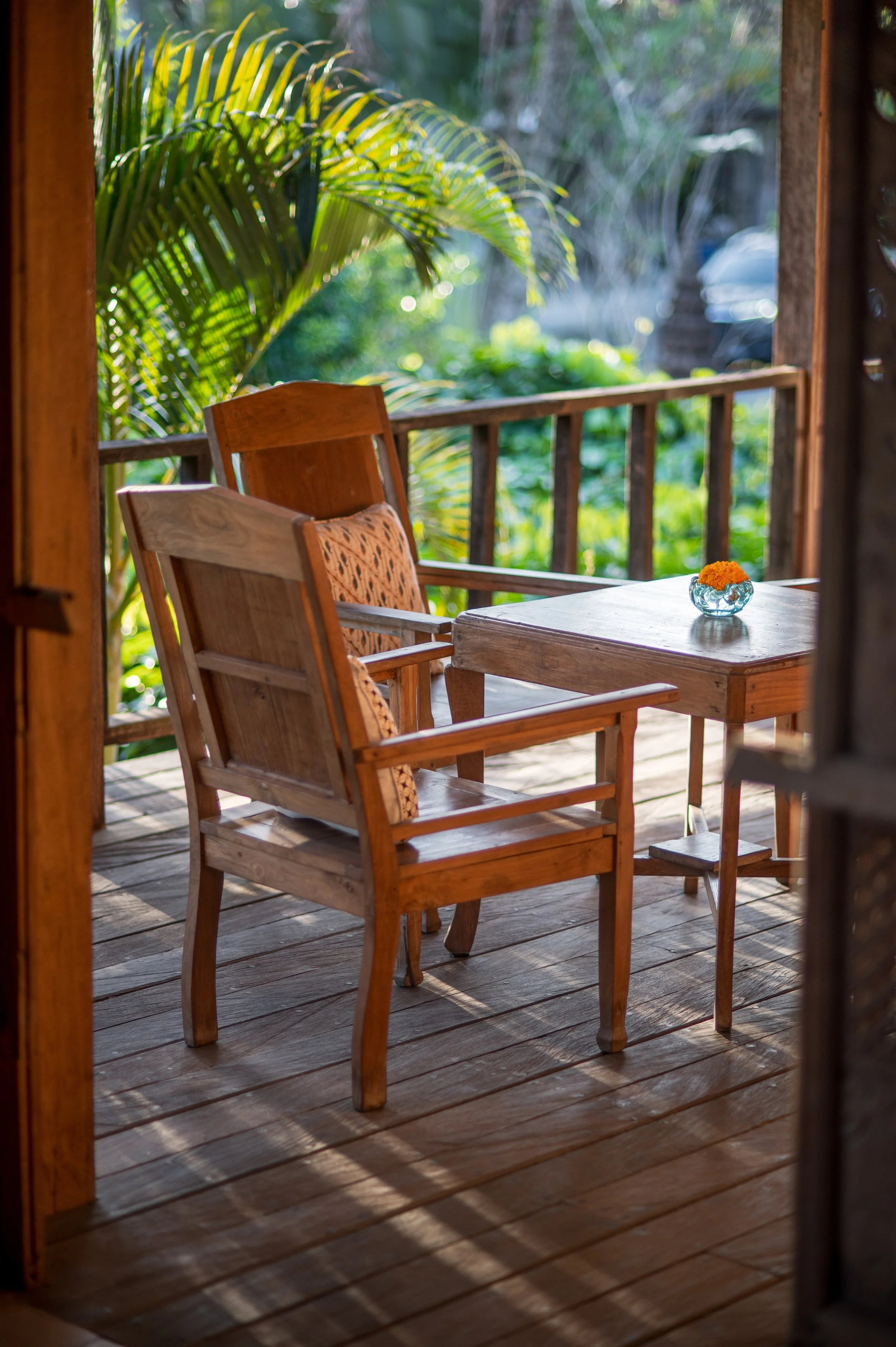 A cozy outdoor wooden porch with two wooden chairs, a small table with a flower in a vase, and lush greenery in the background.