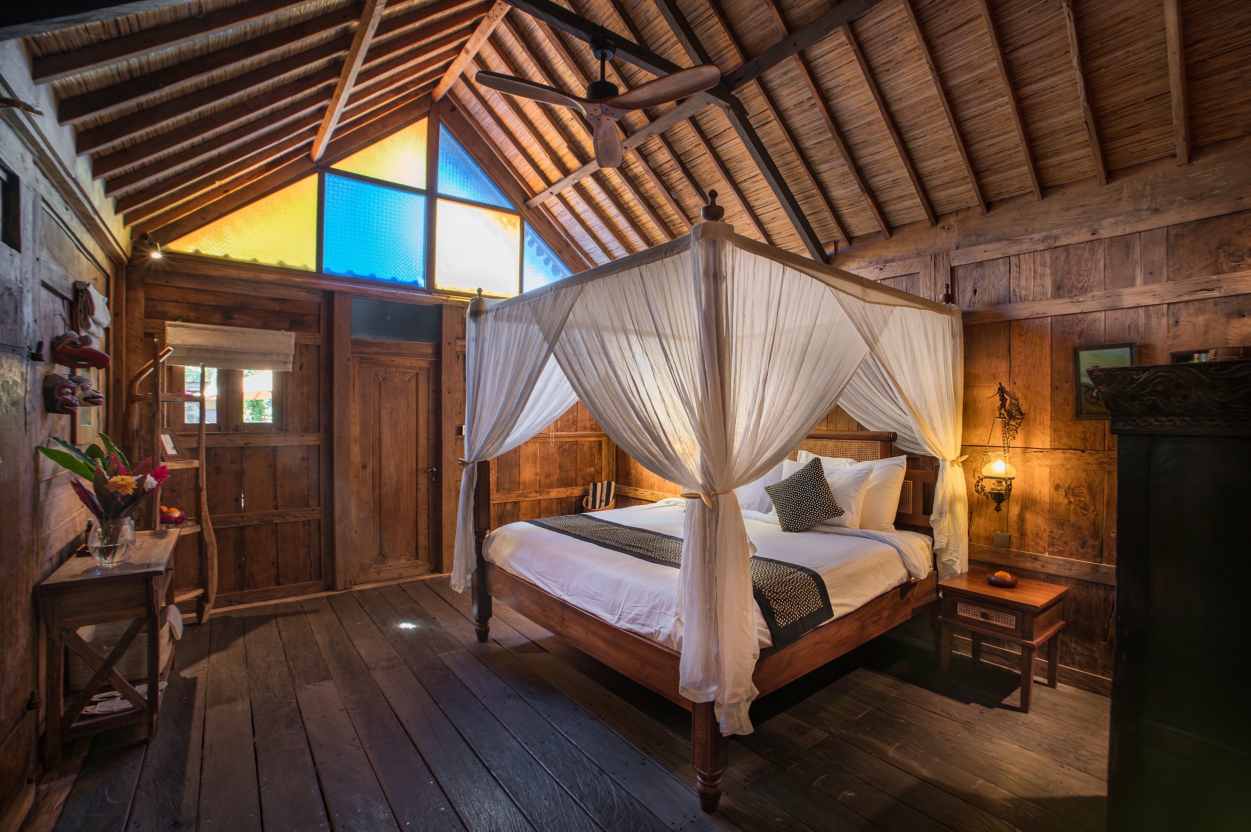 A cozy bedroom with wooden walls and ceiling, featuring a large bed with white linens and mosquito netting, colorful stained glass windows, and warm lighting.