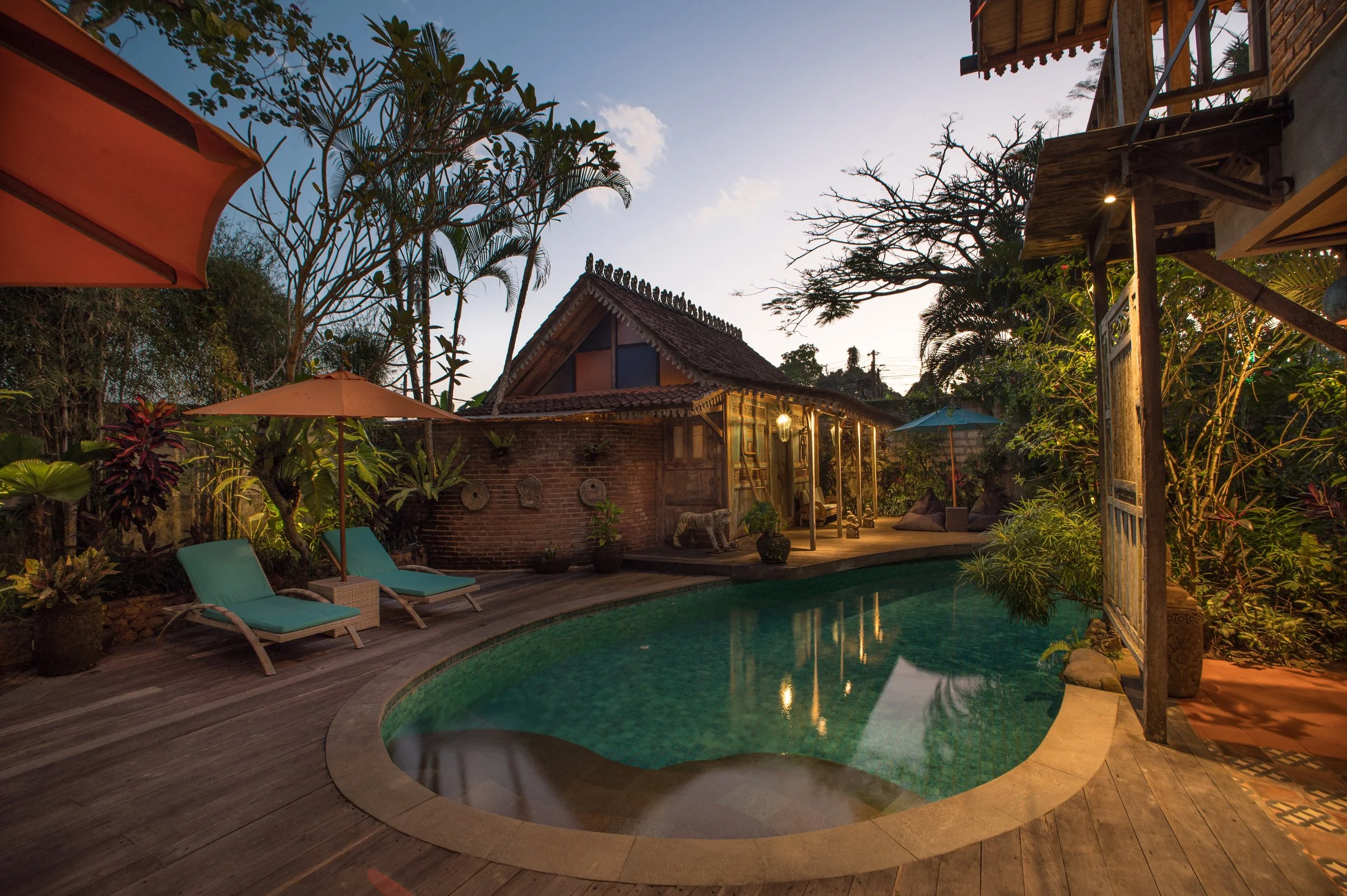 A backyard scene at dusk with a small swimming pool, wooden deck, lounge chairs, umbrellas, tropical plants, and a house with a thatched roof.