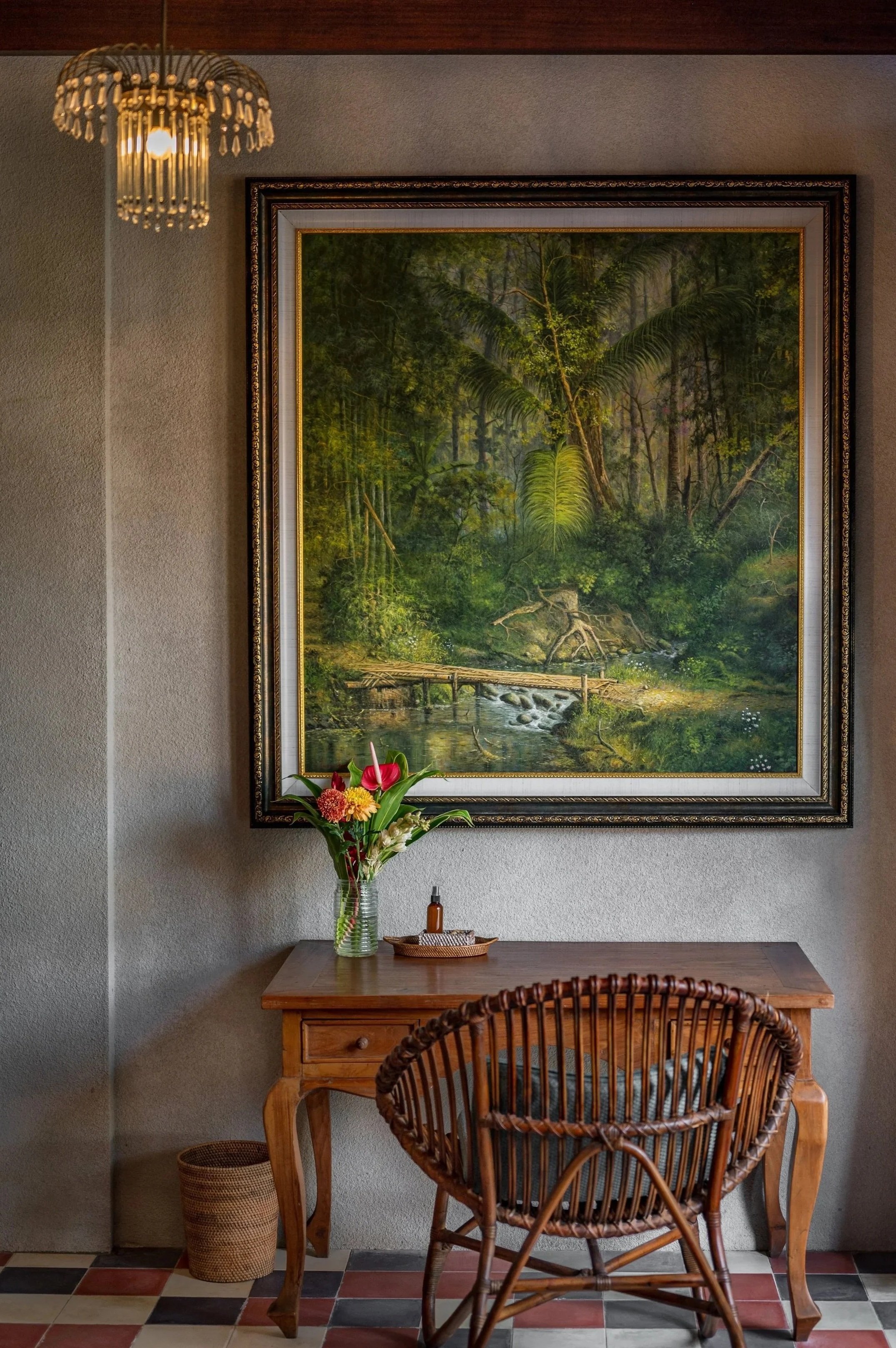 A wooden table with a glass vase of colorful flowers, a small basket, and a bottle of hand sanitizer. Behind the table is a framed landscape painting of a lush forest stream, and a wicker chair is in the foreground. A small wastebasket is beside the table on a tiled floor.