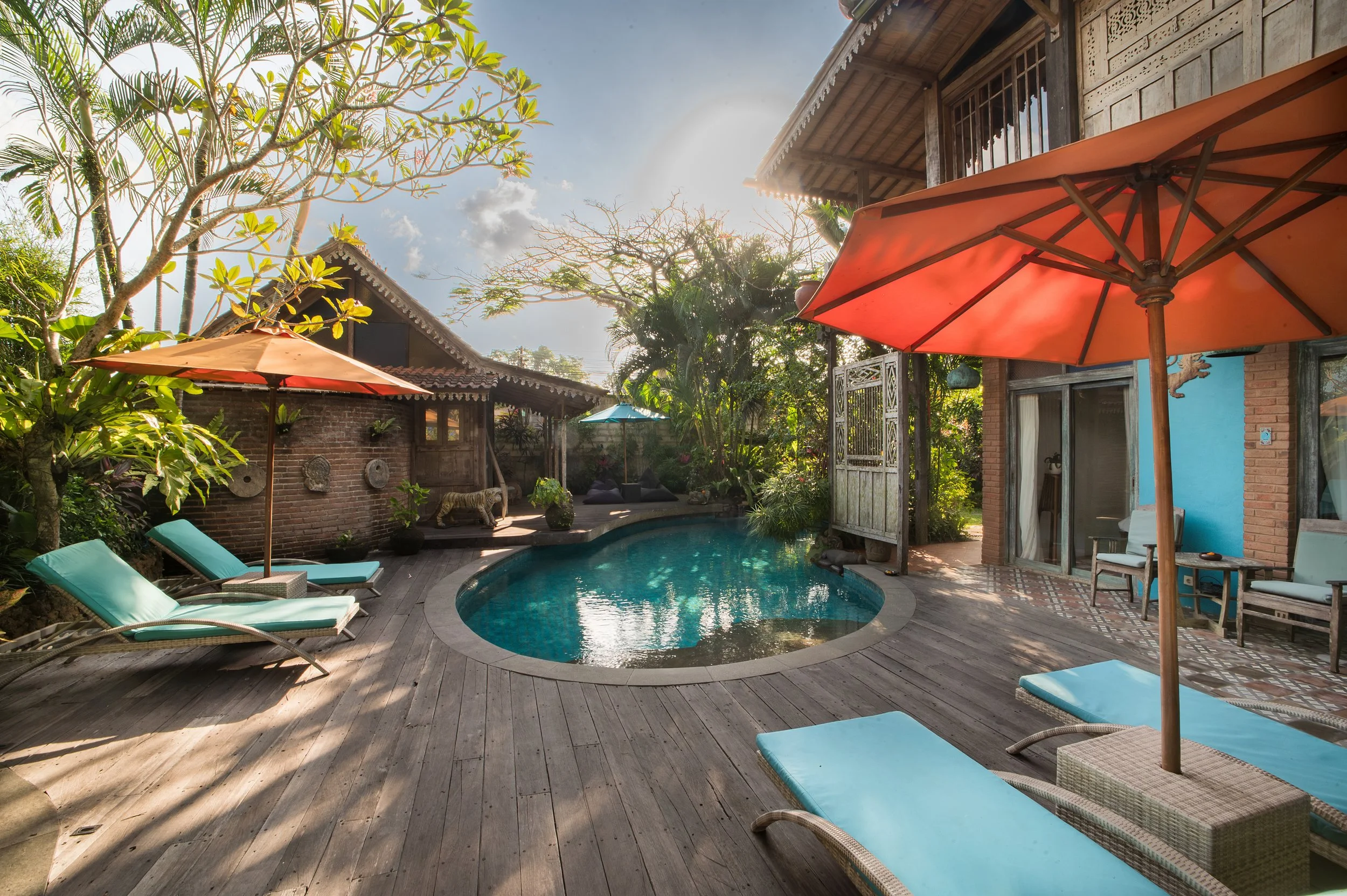 A backyard with a pool surrounded by wooden decking, several lounge chairs with teal cushions, orange patio umbrellas, tropical plants, and a traditional style house with wooden and brick elements, under a partly cloudy sky with the sun shining.
