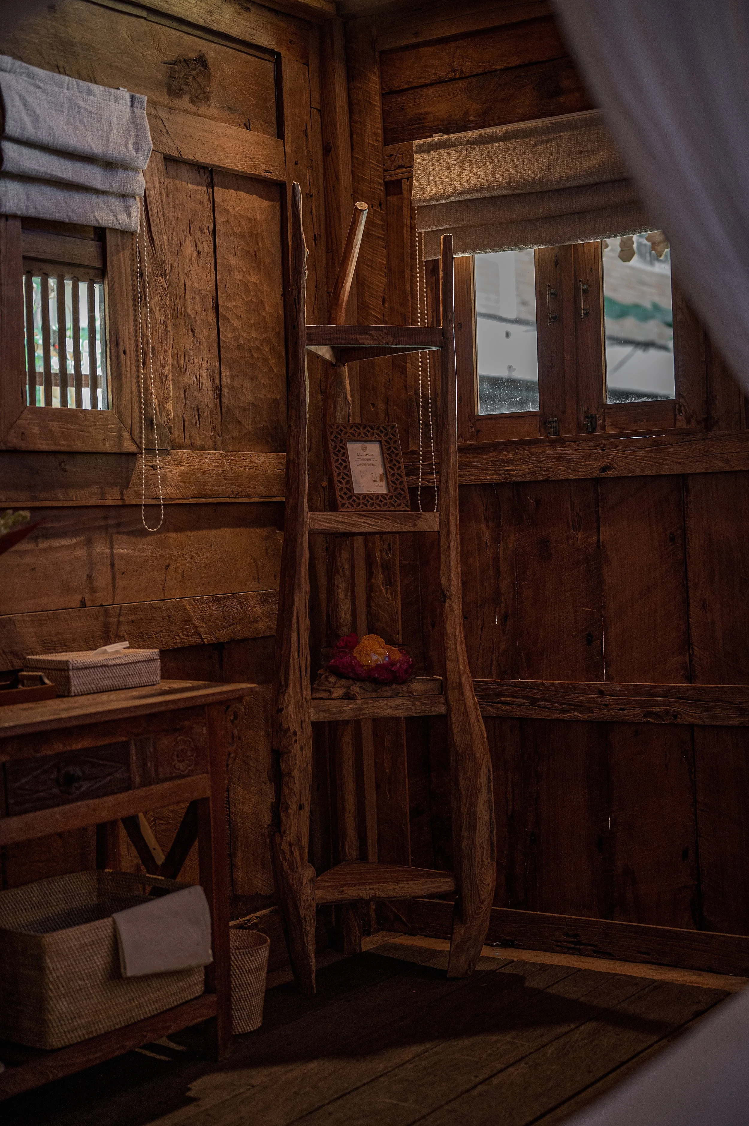 Interior of a rustic wooden cabin with a wooden ladder, window with fabric blinds, framed picture, and a small table with a basket.