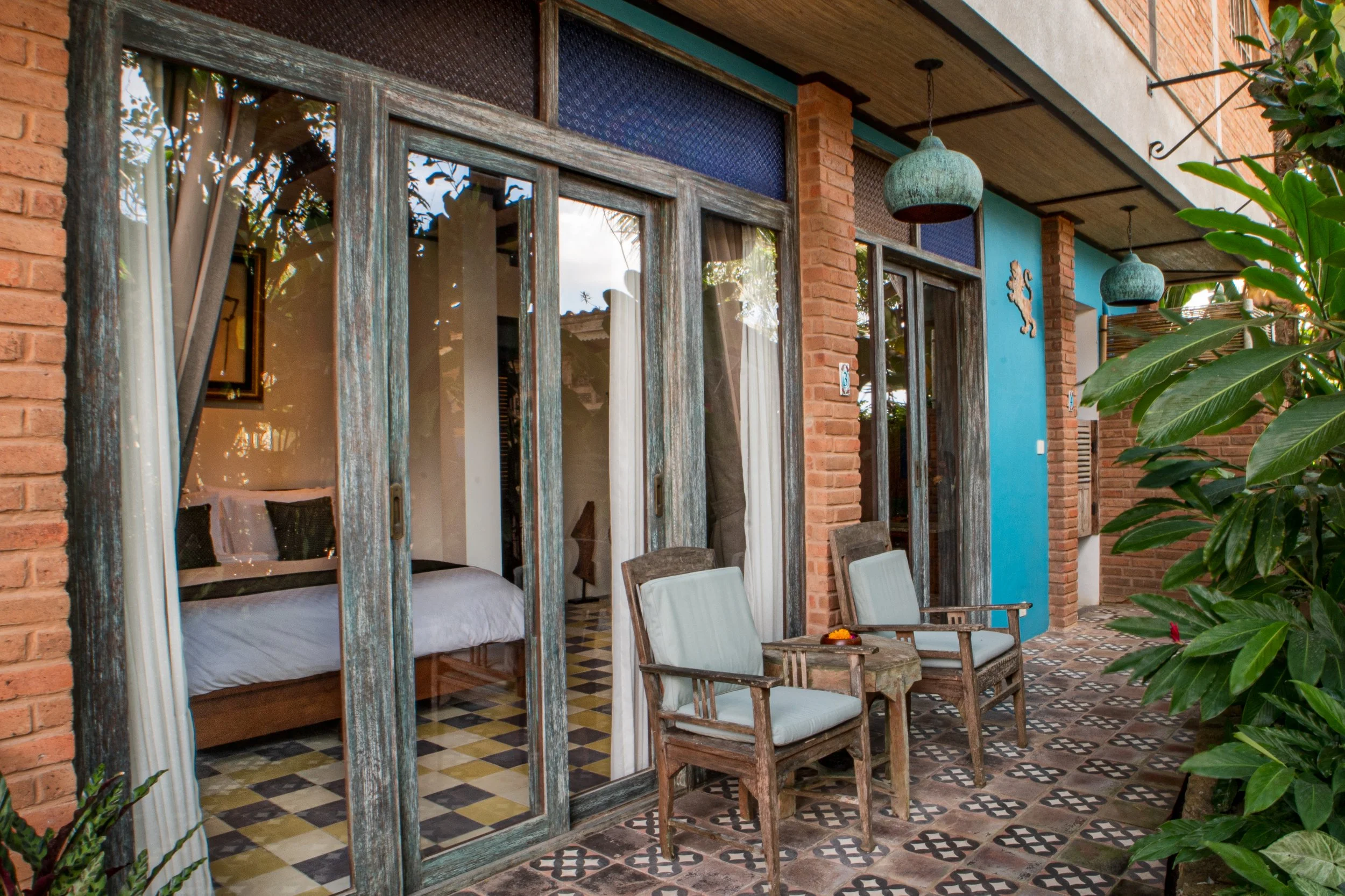 A patio area with wooden chairs and a small table outside a house with large glass sliding doors, brick walls, and hanging teal lamps, surrounded by green plants.
