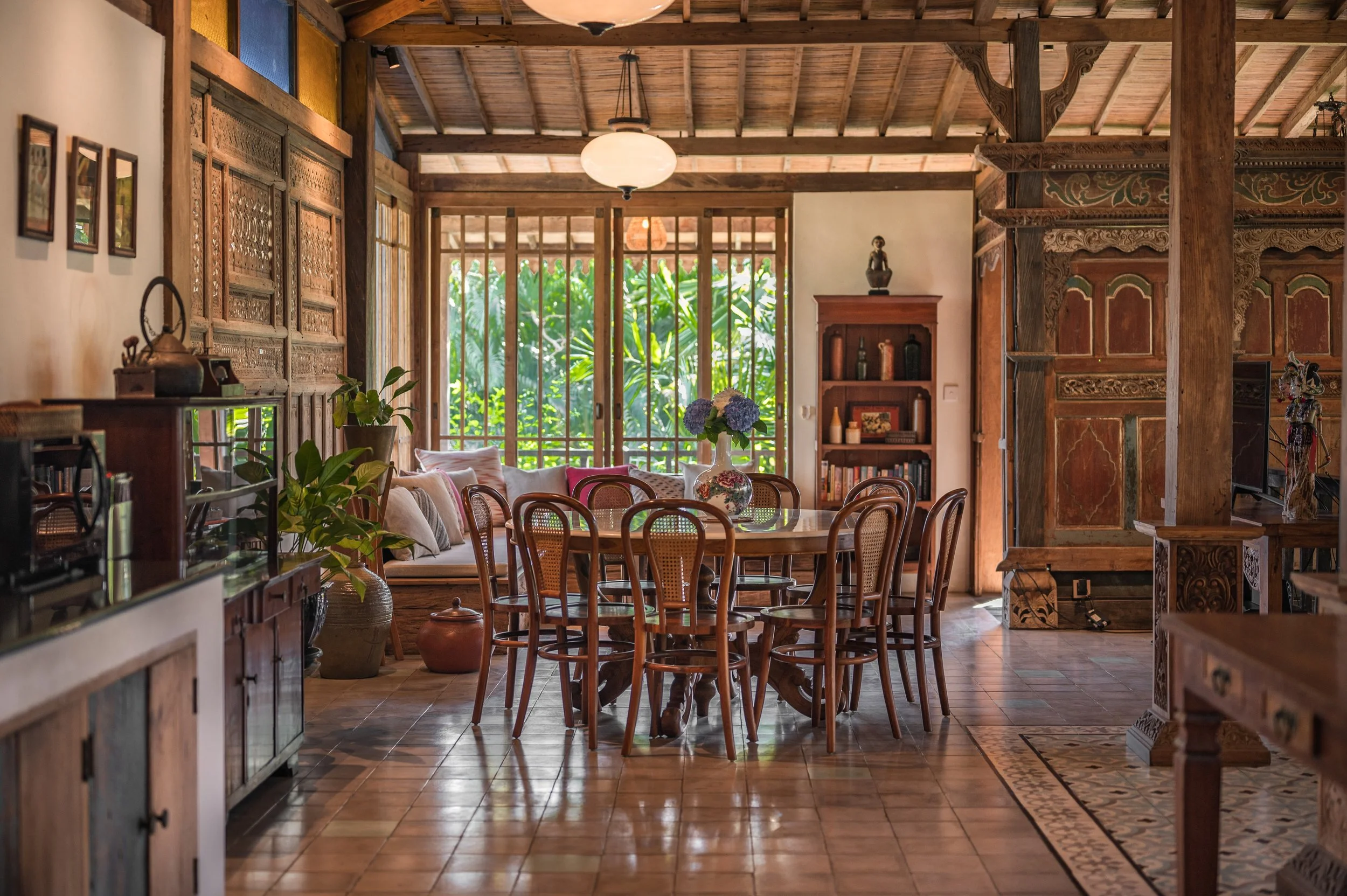 A cozy dining and living room area with wooden furniture, a central round table with chairs, a large floral vase, and a window with greenery outside. The room features wooden beams, carved wood panels, and a tiled floor, creating a warm and inviting 