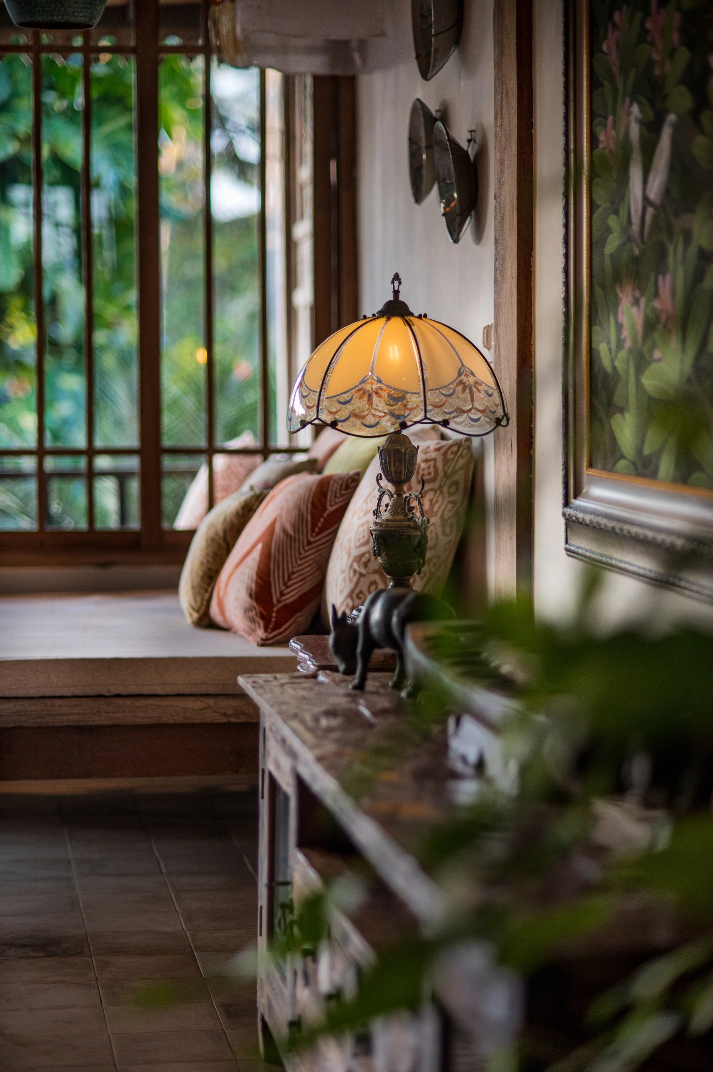 Inside a cozy living space with a vintage stained glass lamp on a wooden table, cushions on a step, and a large window with wooden framing showing greenery outside.