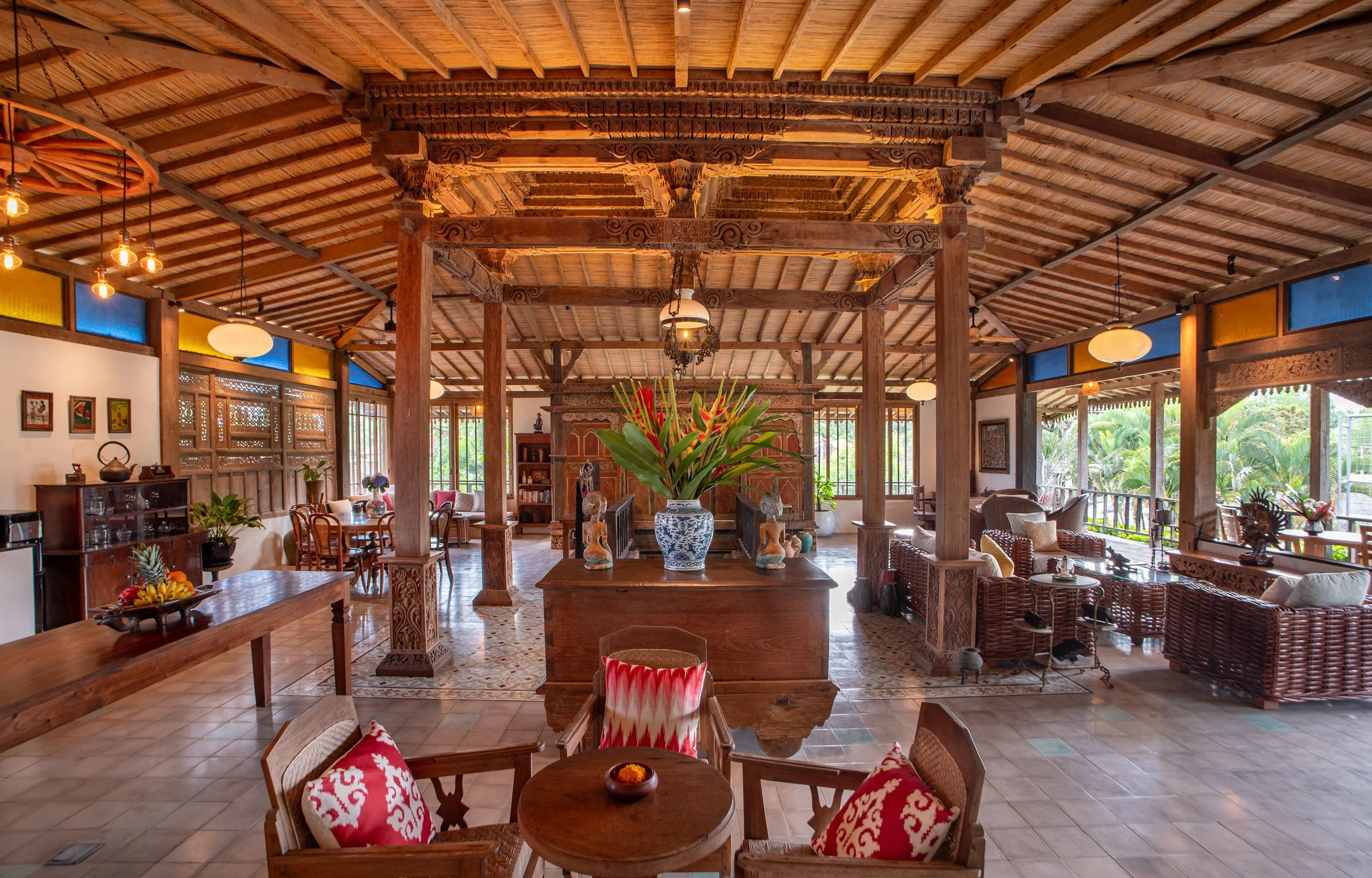 Spacious interior of a house with wooden beams and furniture, potted plants, colorful walls, and large windows showing greenery outside.