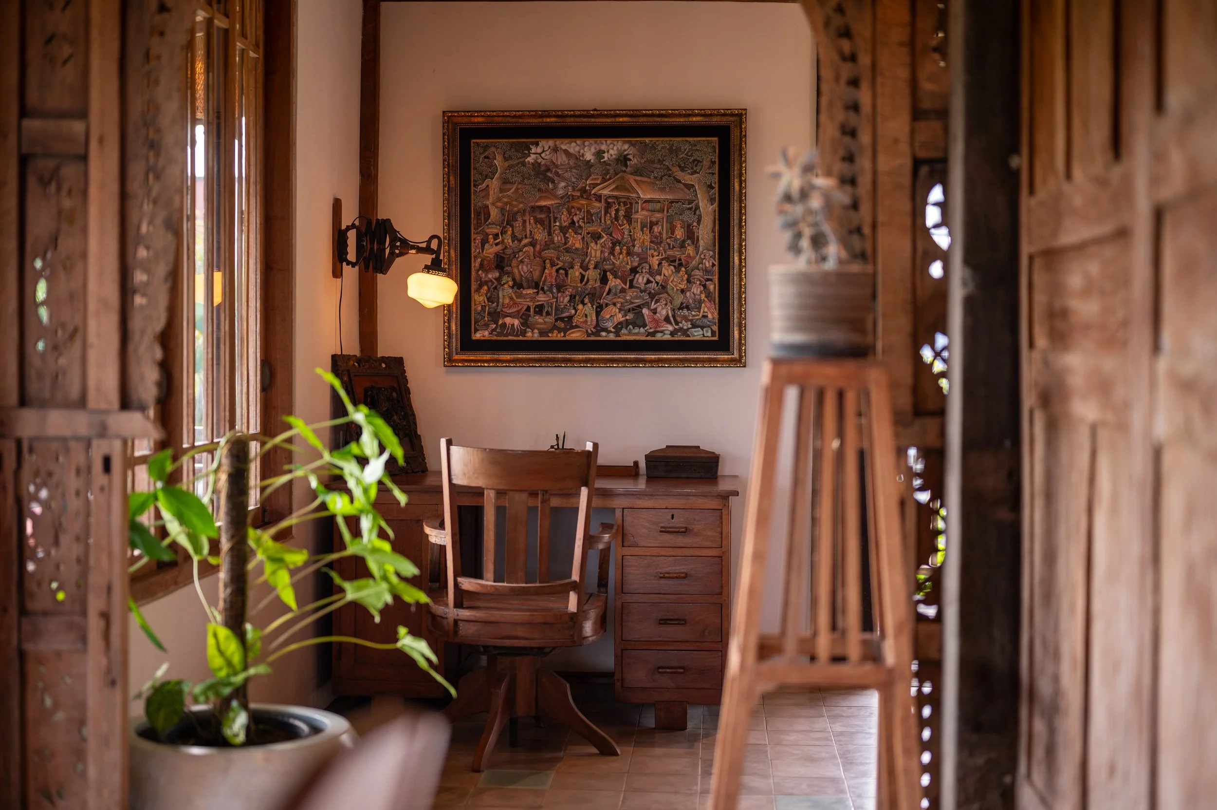 A cozy wooden study or home office with a wooden desk, chair, a framed artwork on the wall, a small table, a wall-mounted lamp, and a potted plant, seen through wooden slats or room divider.