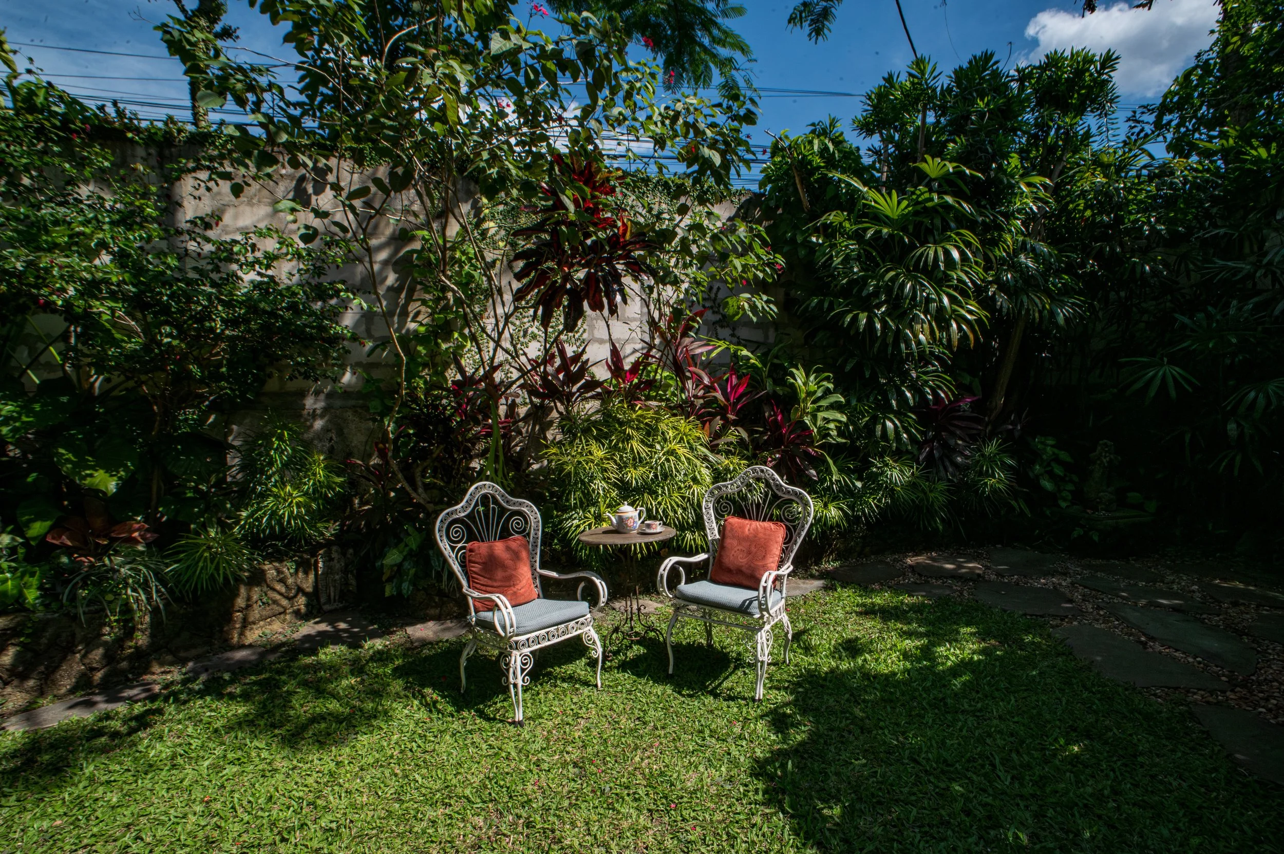 A cozy outdoor seating area with a small round table and two ornate metal chairs with orange cushions, surrounded by lush greenery including trees and bushes, under a partly cloudy sky.