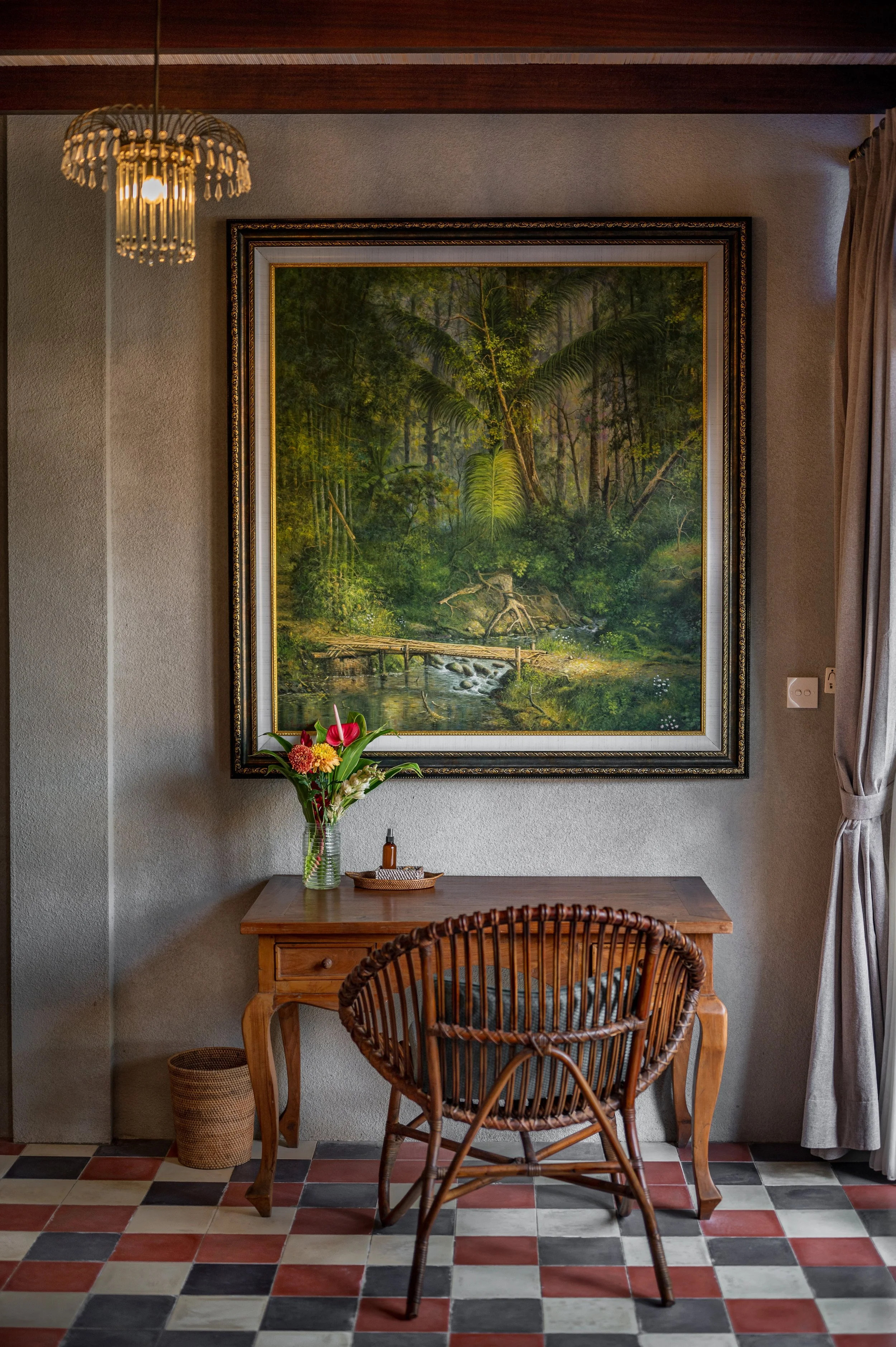 A room with a wooden desk, a wicker chair, and a colorful tiled floor. A large framed painting of a lush green forest scene hangs on the wall. On the desk, there is a glass vase with vibrant flowers, a small tray, and a bottle. A curtain is partially