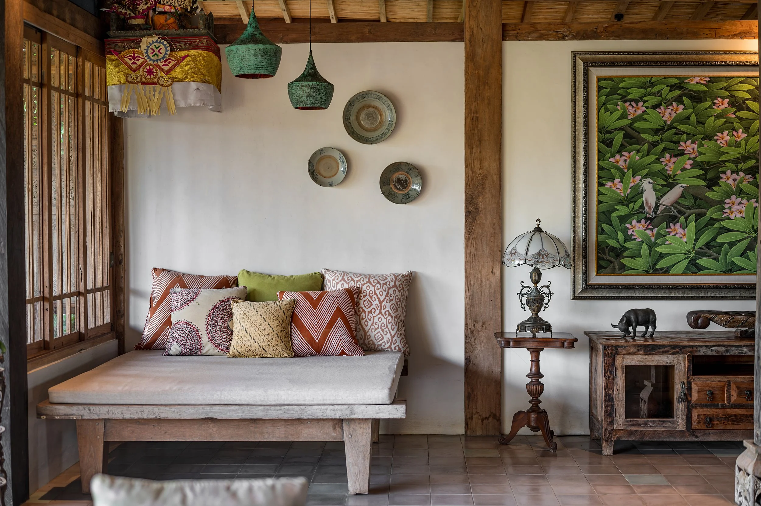 A cozy interior corner with a wooden bed frame and an assortment of patterned pillows, a side table with a vintage lamp, decorative plates on the wall, and framed lush green foliage artwork.