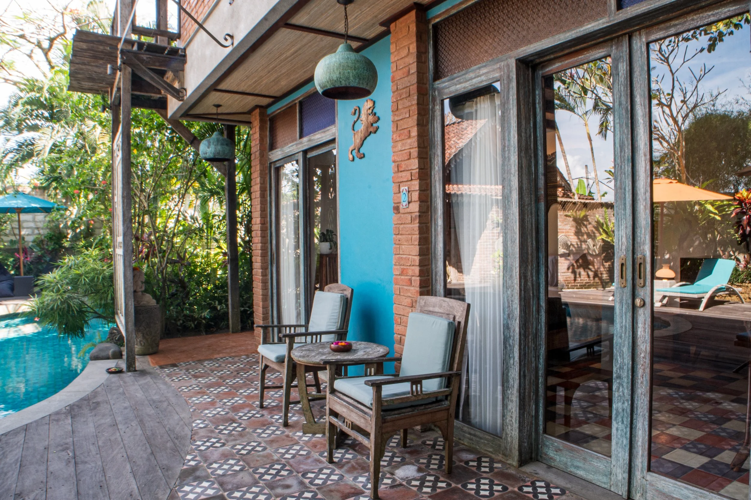 View of a patio with outdoor seating, a swimming pool, lush greenery, and a house with large glass sliding doors and a blue wall decorated with a lion ornament.