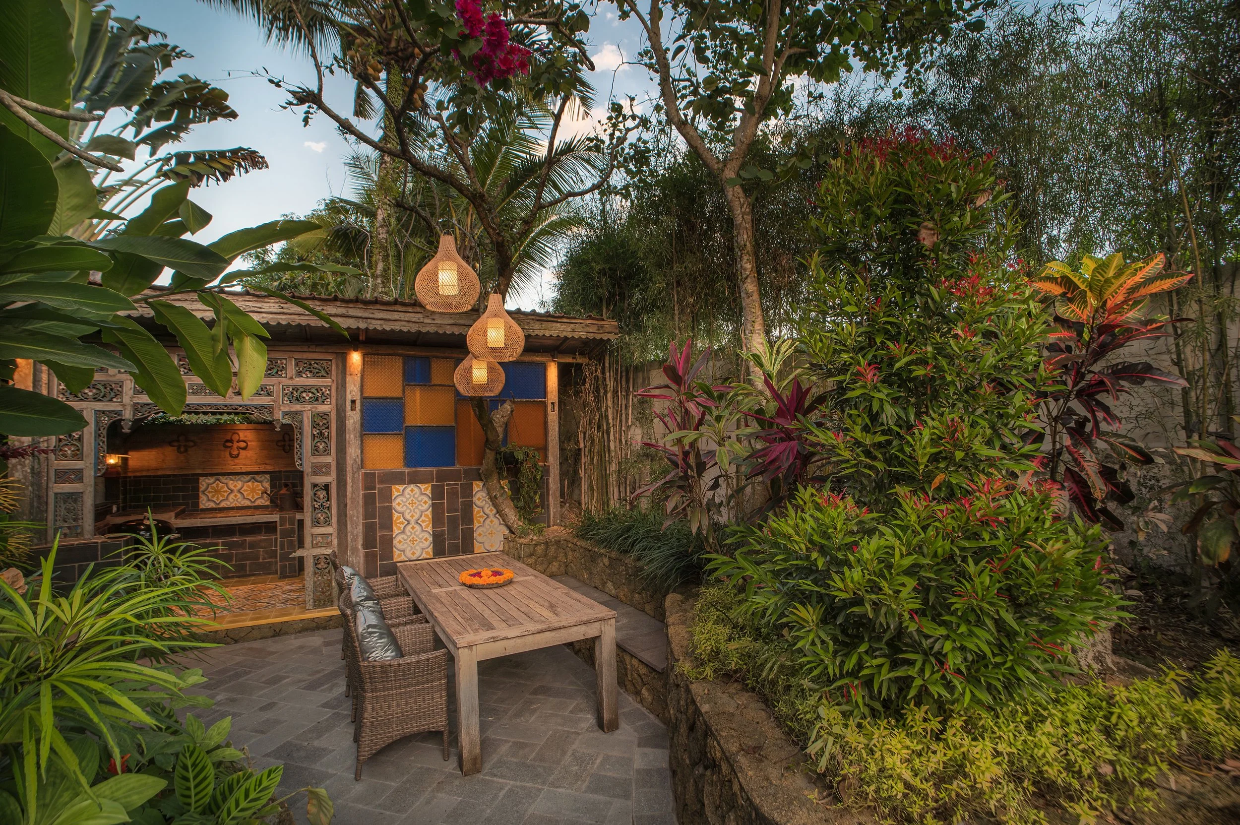 Outdoor patio with wooden dining table and wicker chairs surrounded by lush green plants and trees, with decorative hanging lanterns and a colorful tiled wall in the background.