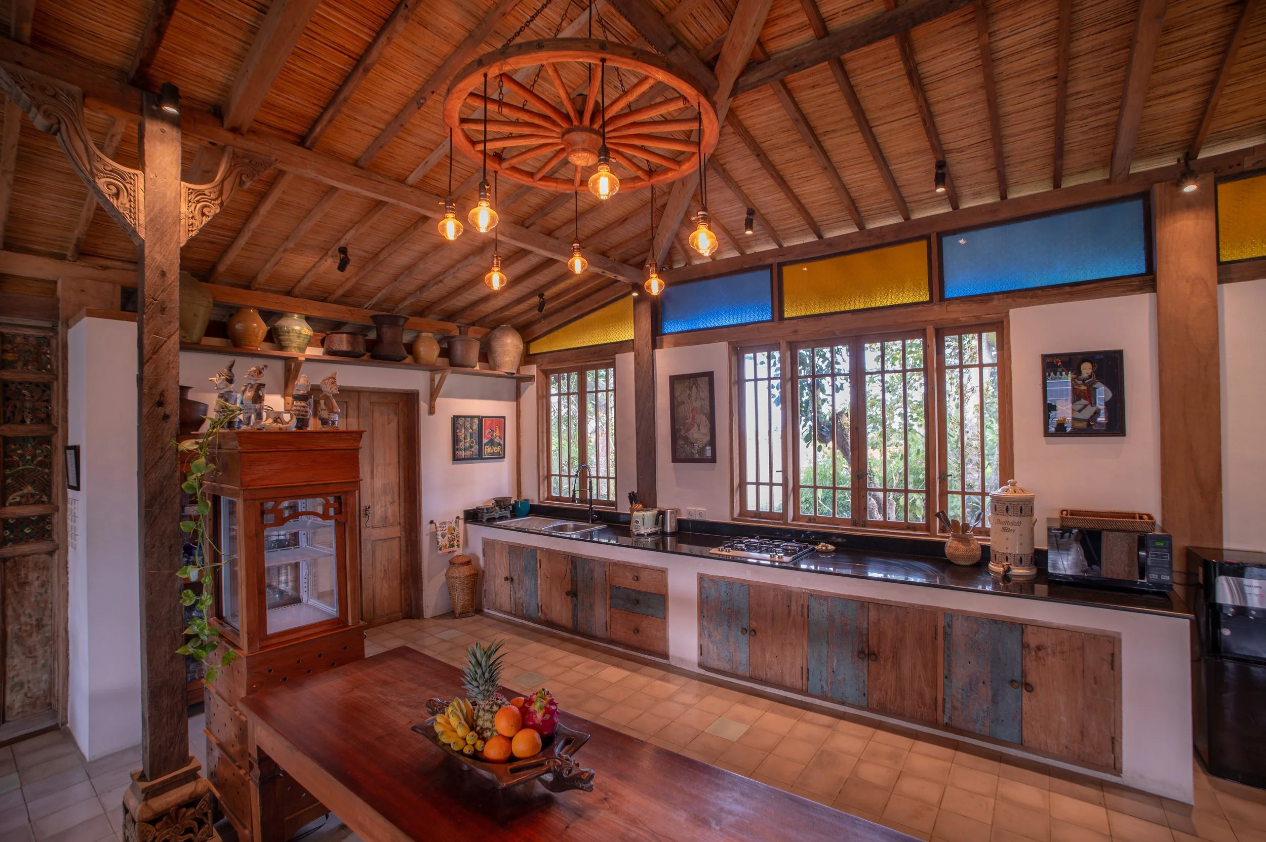 Rustic kitchen with wooden ceiling, large window, pendant lights hanging from a wagon wheel chandelier, and shelves with clay pots. There is a wooden dining table with a fruit bowl, and various kitchen appliances and decor.