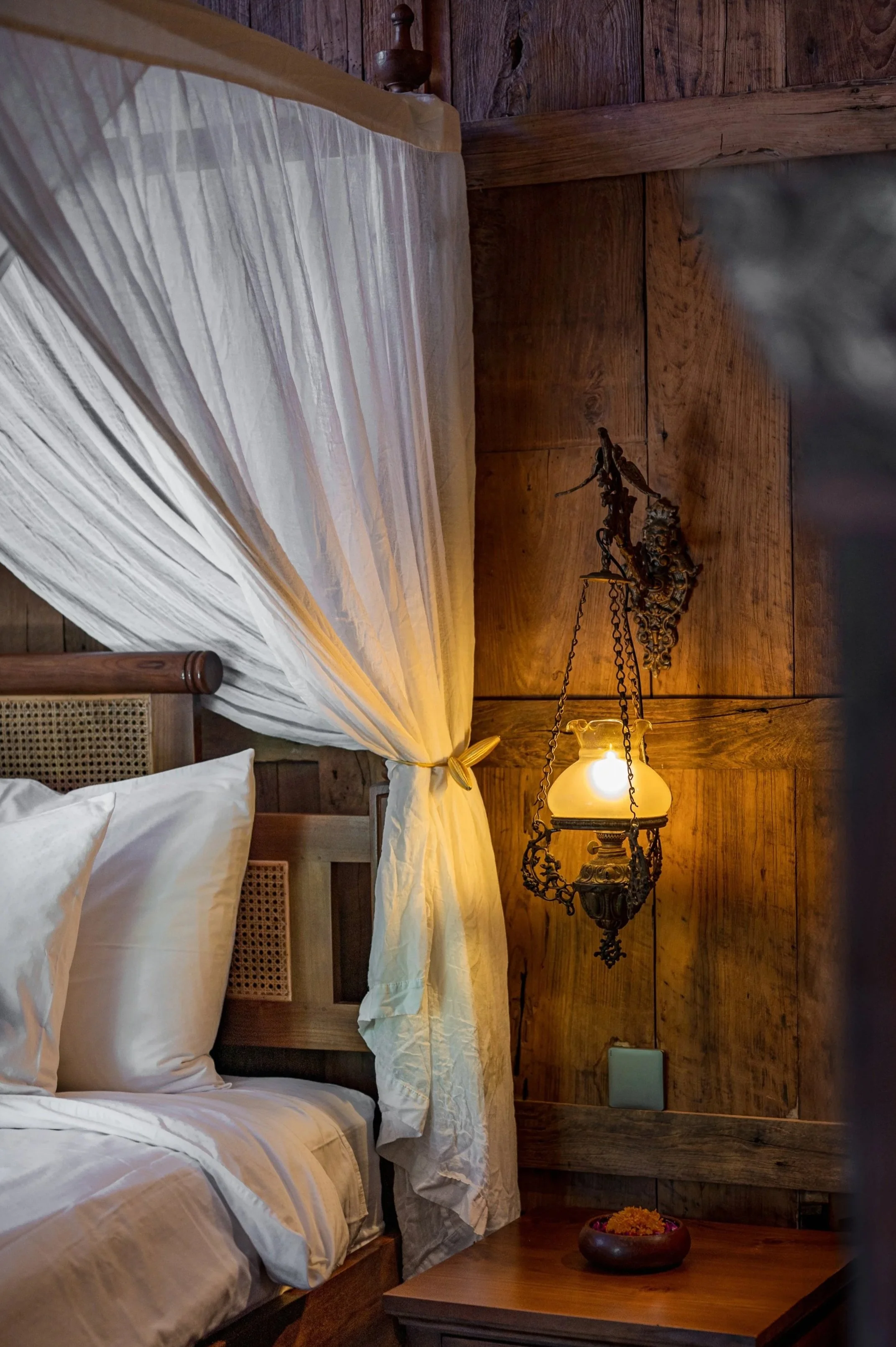 A bedroom with wooden paneled walls, a bed with white linens and a canopy, a wooden bedside table with a small bowl of orange flowers, and an ornate hanging lamp with a warm light.