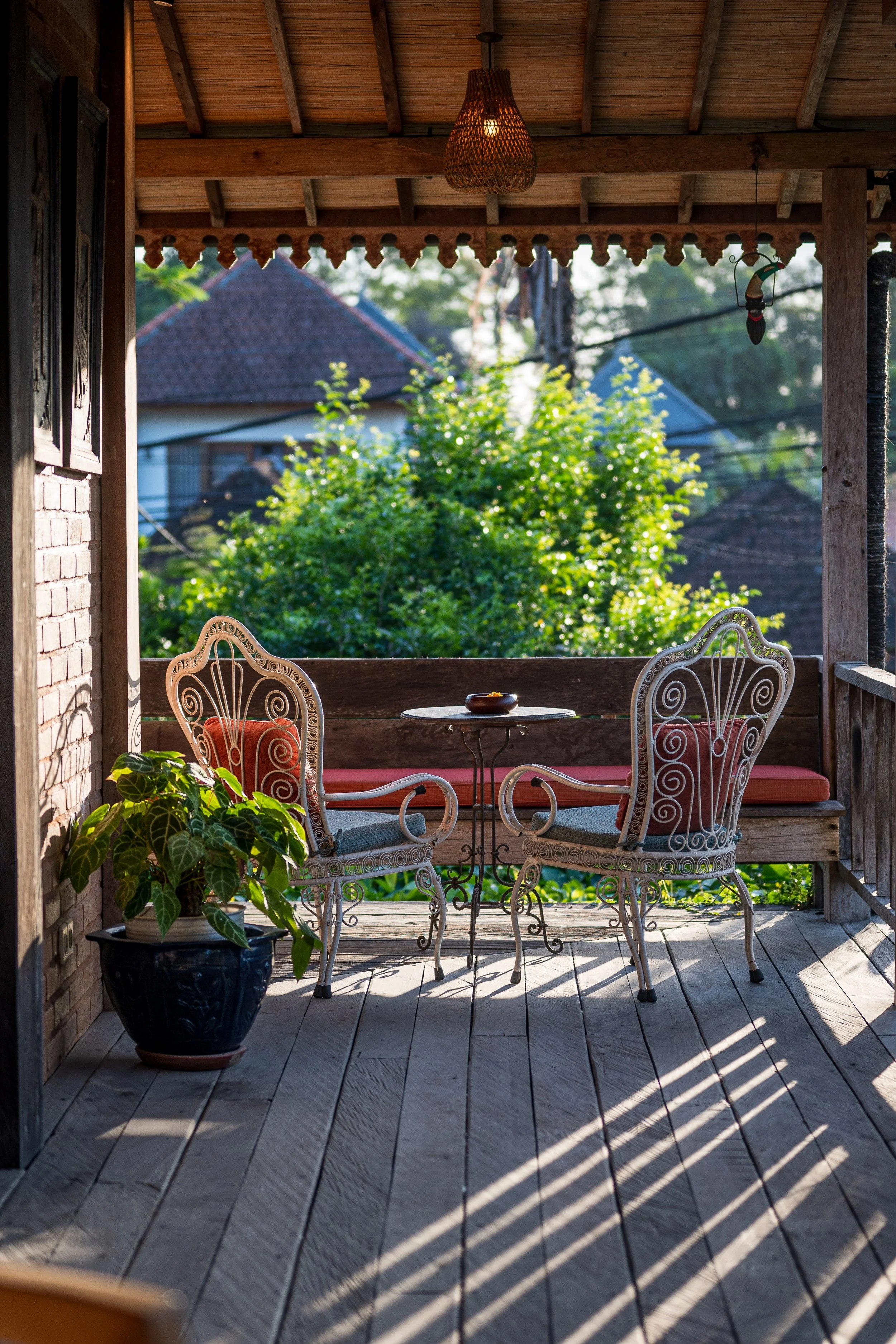 A cozy outdoor balcony with white wrought iron chairs, a small round table, a large potted plant, and lush greenery in the background, sunlit with shadows cast on the wooden floor.