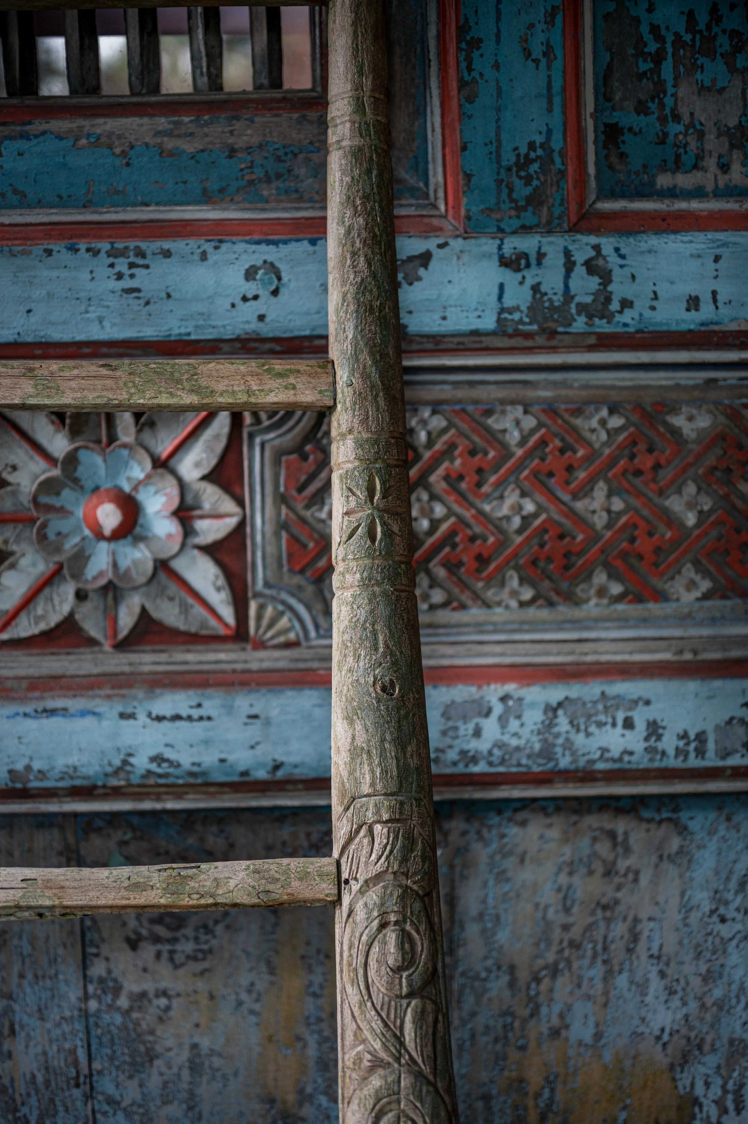Close-up view of a weathered wooden pole with carved patterns, standing in front of an ornate, distressed painted ceiling with floral and geometric designs in blue, red, and white.