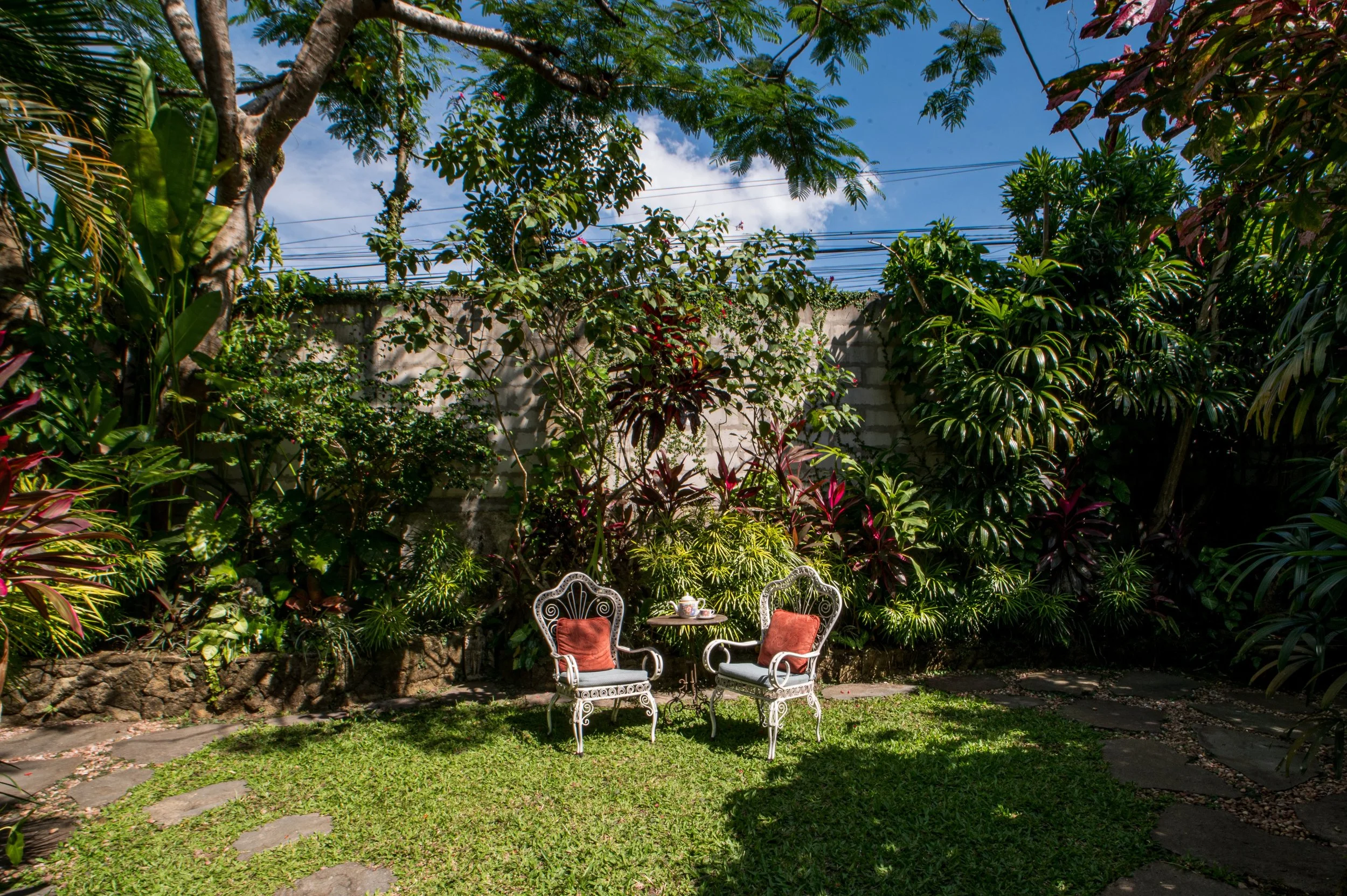 A lush garden with green plants and trees, a stone wall in the background, blue sky with clouds, and three white wrought-iron chairs with red cushions and a small white teapot and cup on a table.