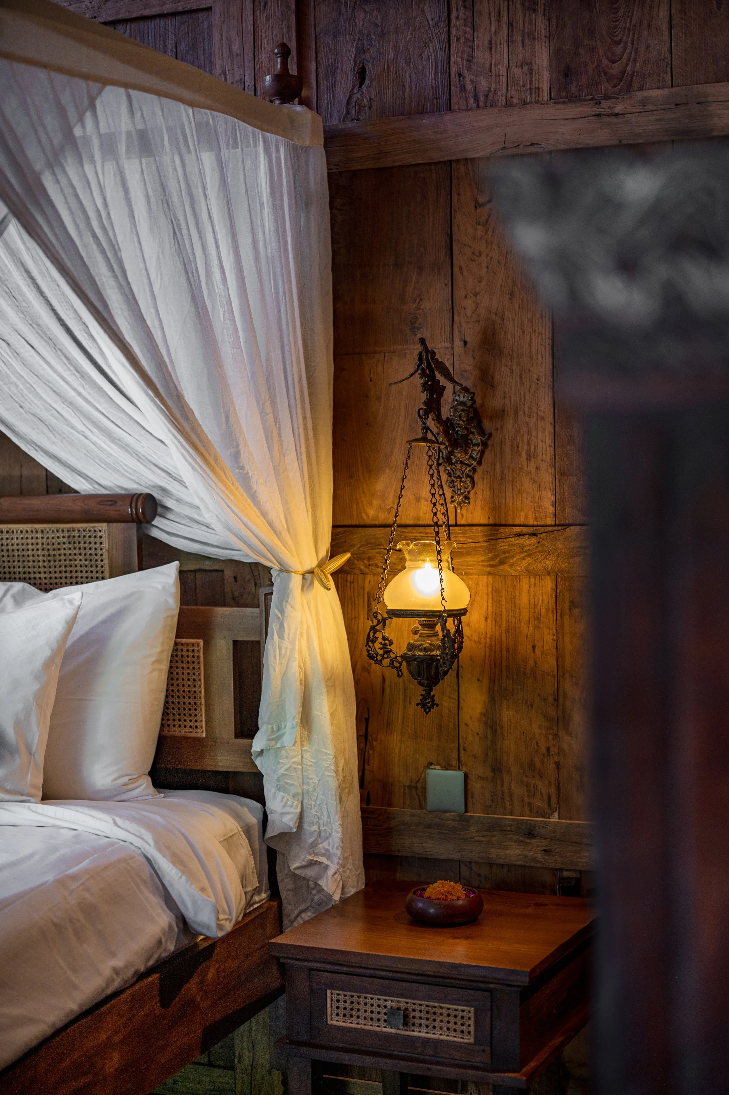 A rustic bedroom with wooden walls, a window with white curtains, a bed with white bedding, a small bedside table with a bowl of flowers, and a vintage hanging lamp.