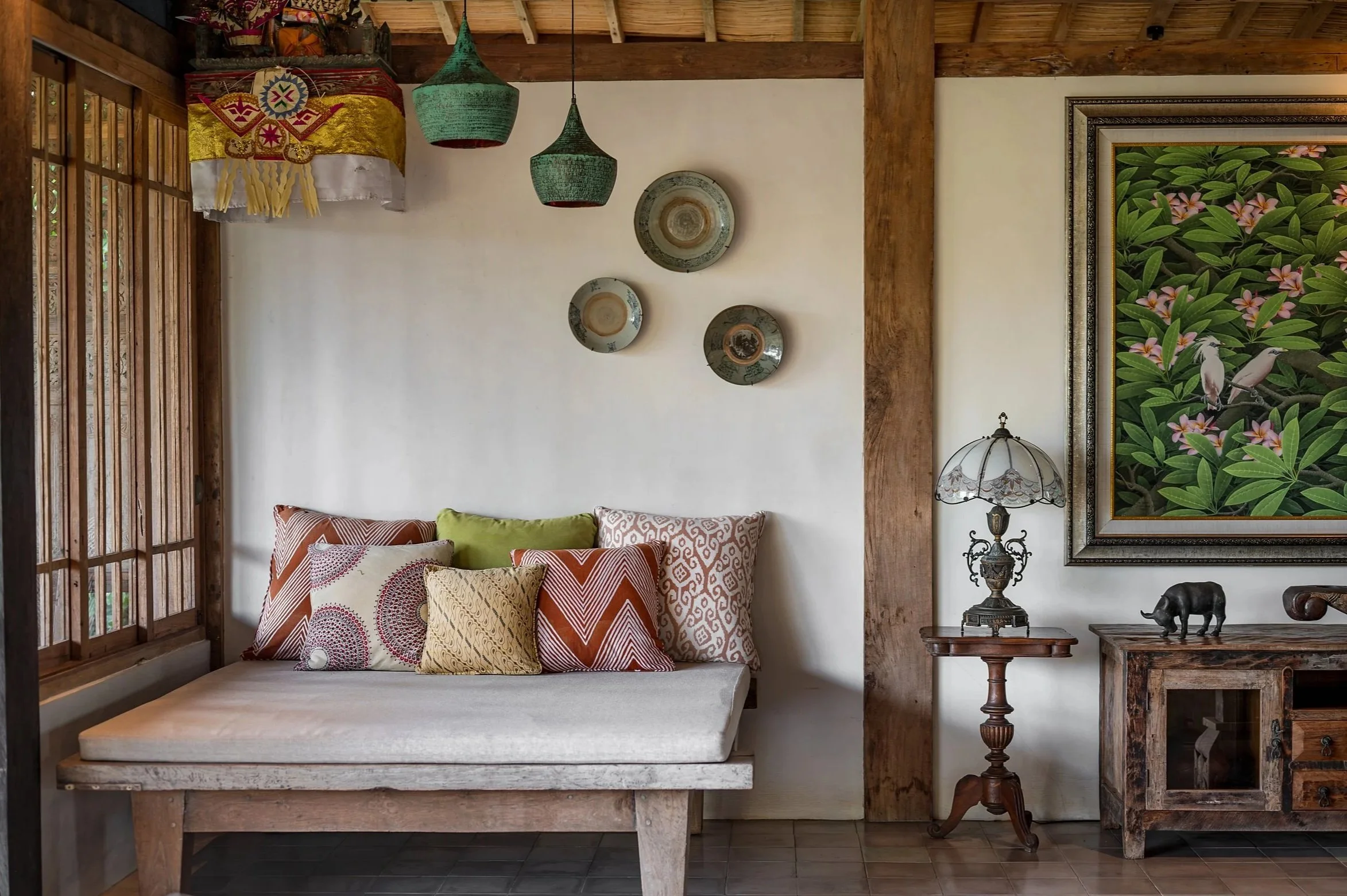 A cozy corner of a living room with a wooden ceiling and paneling, featuring a large white wall decorated with four ceramic plates and two hanging green woven lamps. A colorful framed painting of birds on a leafy background hangs on the right. There is a light-colored sofa with various patterned throw pillows, a carved wooden side table with a vintage lamp, and a wooden figurine of an animal on the table.