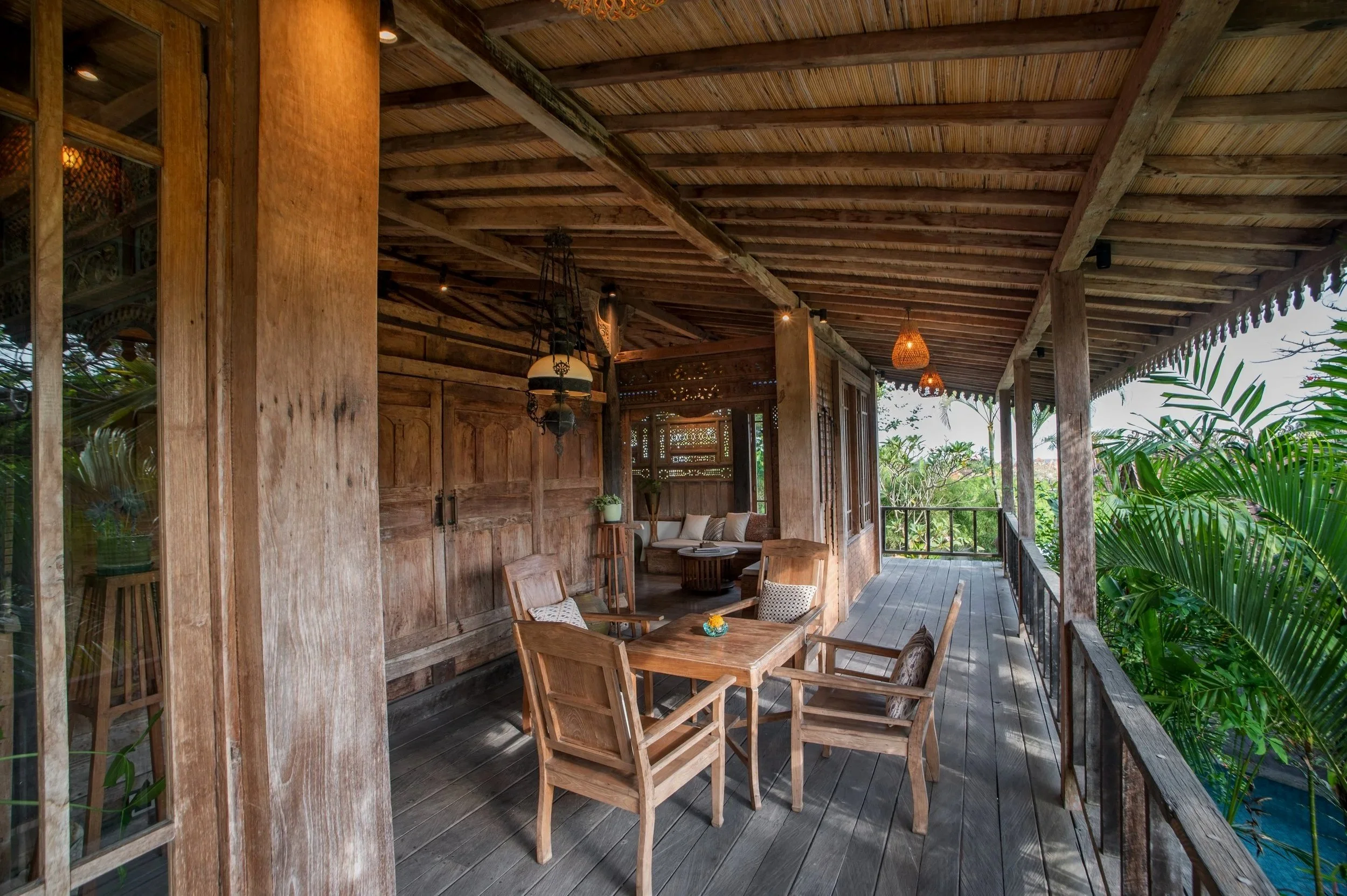A covered wooden balcony with outdoor furniture and hanging lamps, overlooking lush greenery.