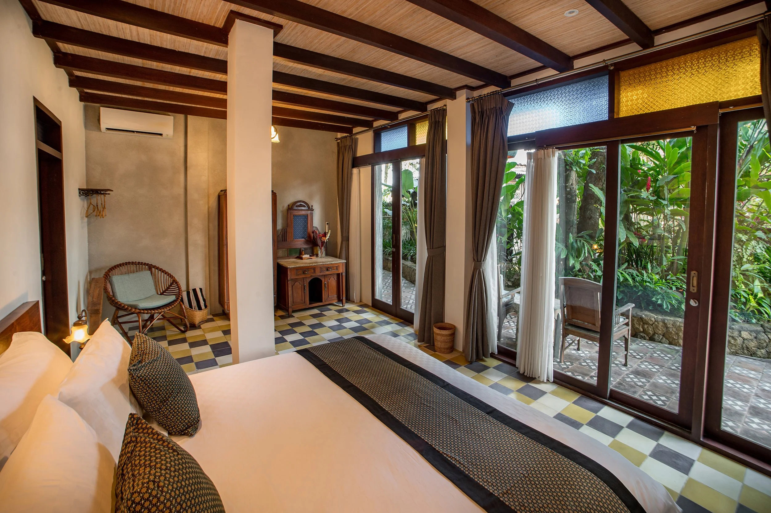 Interior view of a room with large glass doors opening to a lush green garden, featuring a bed with patterned pillows, wooden furniture, and a checkered tile floor.