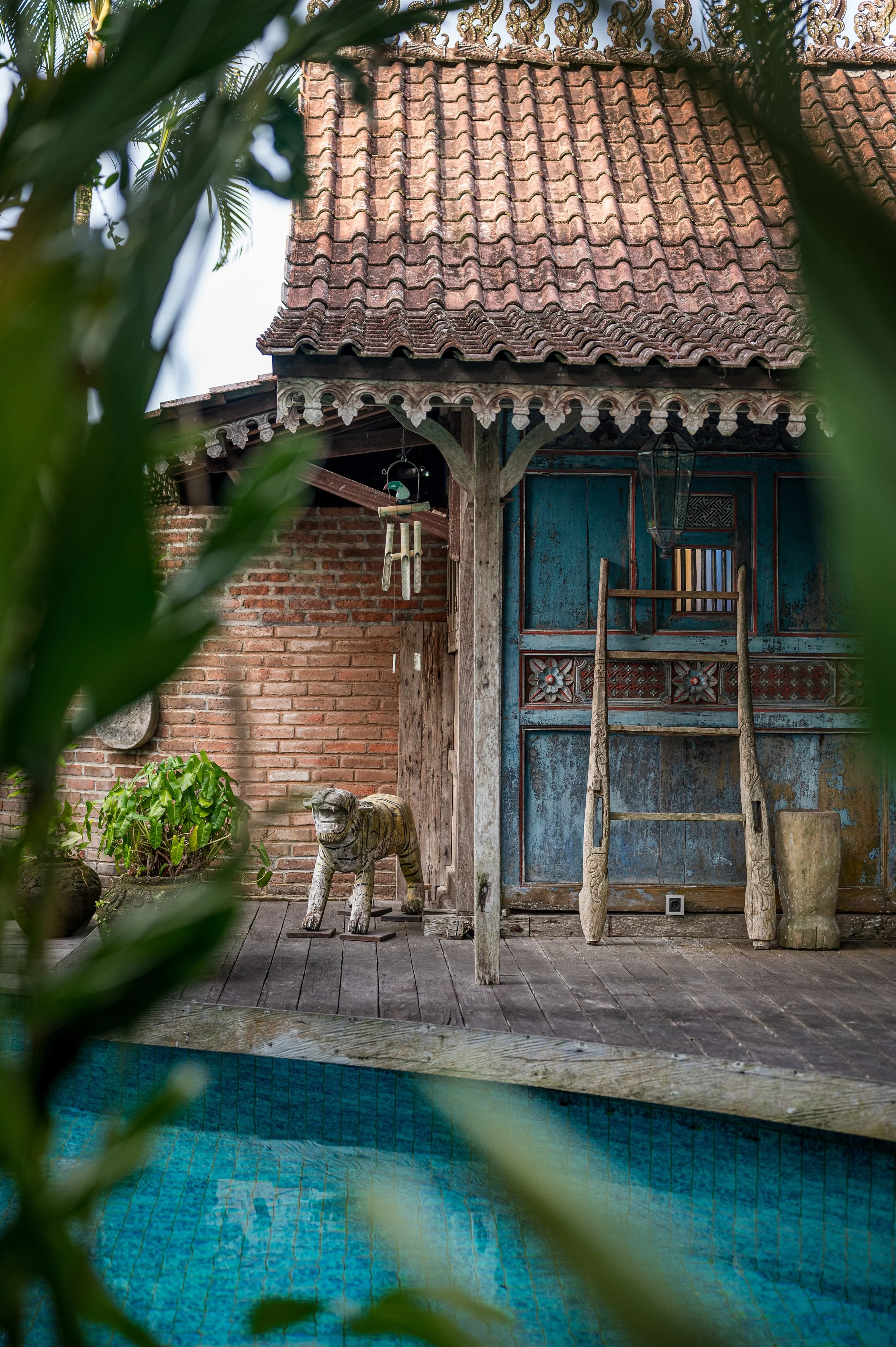 A backyard scene with a small swimming pool in the foreground, surrounded by greenery. In the background, there's a rustic wooden structure with a brick wall, weathered blue painted wall, and a tiled roof. Various decorative items, including a wooden