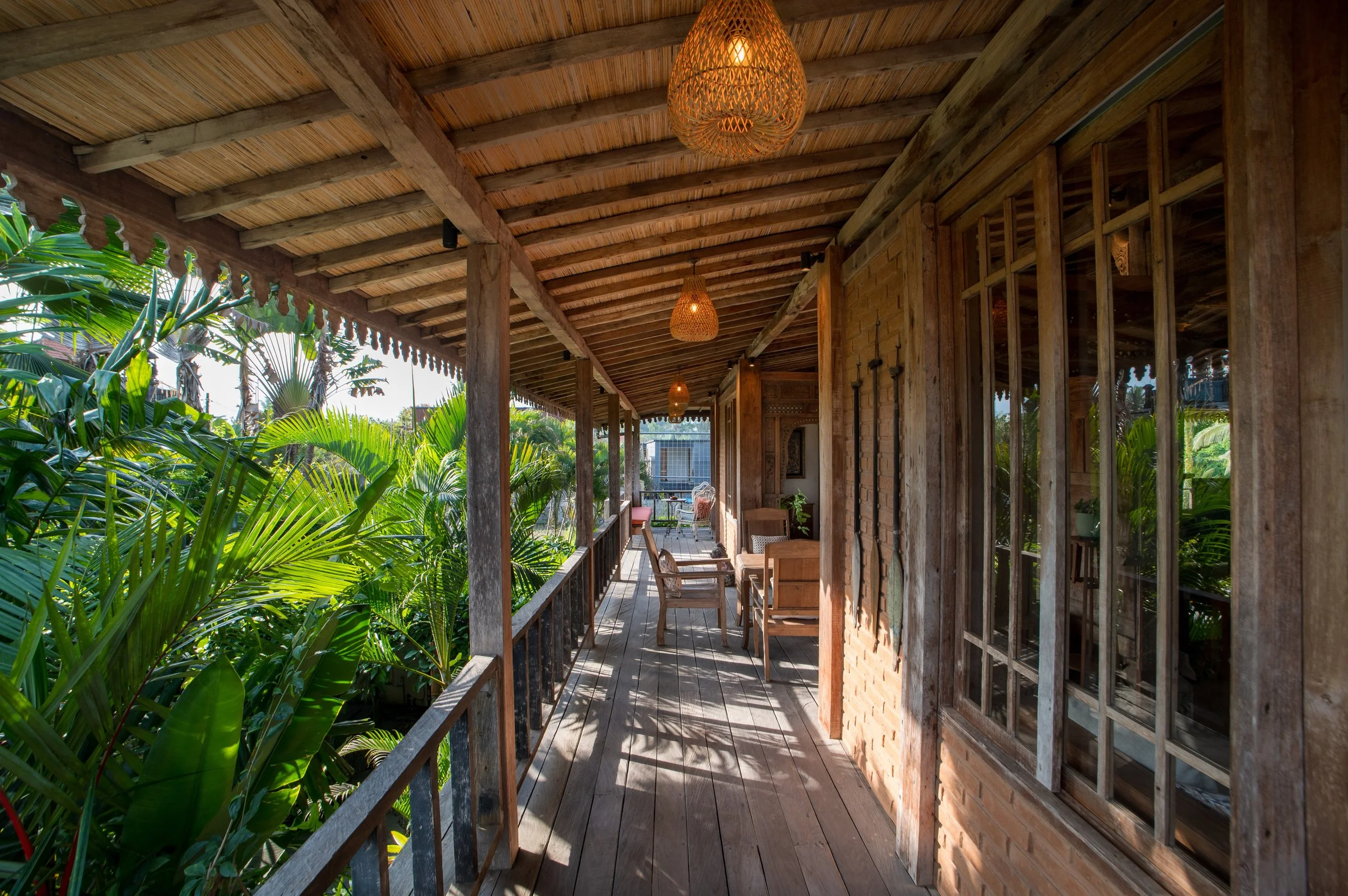 Covered porch with wooden beams and hanging wicker lights, surrounded by lush green tropical plants.