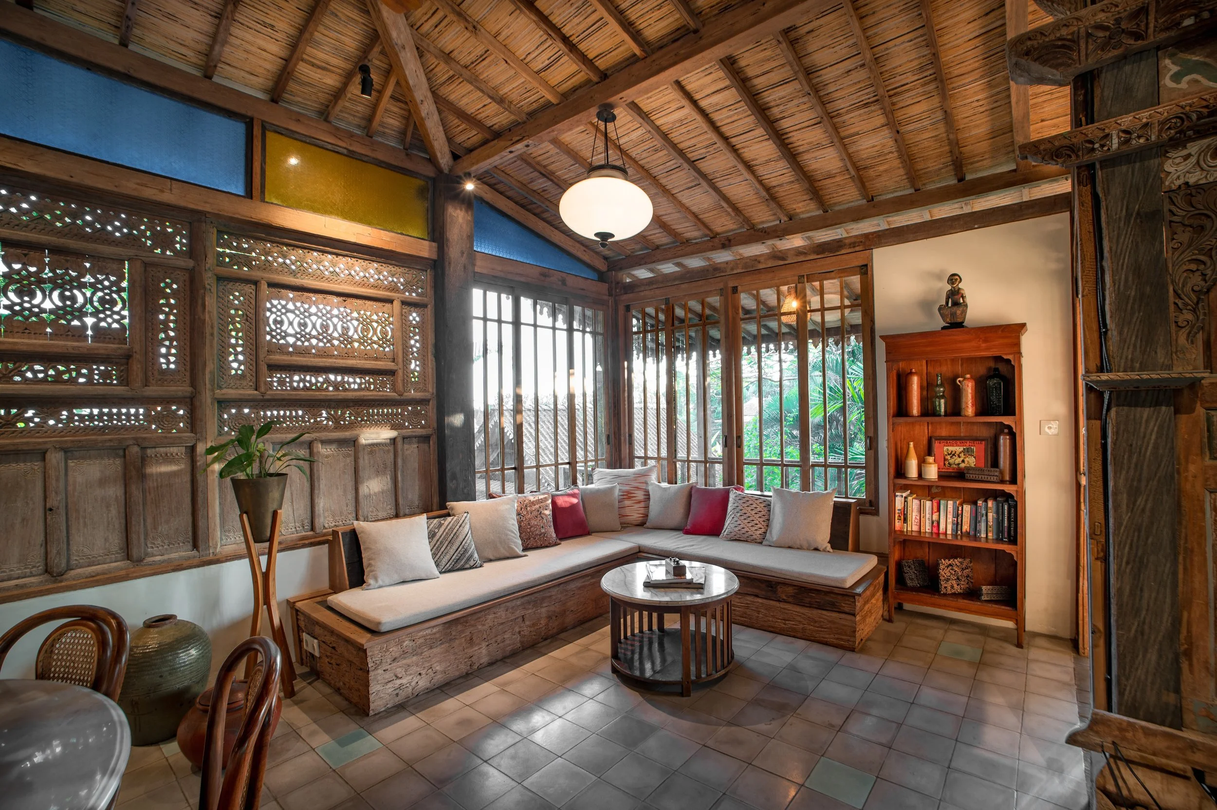 Interior of a cozy room with a high wooden ceiling, large windows, and a built-in wooden sofa with beige cushions and decorative pillows, a small round coffee table, a tall wooden bookshelf with decorative items and books, and some artwork and plants