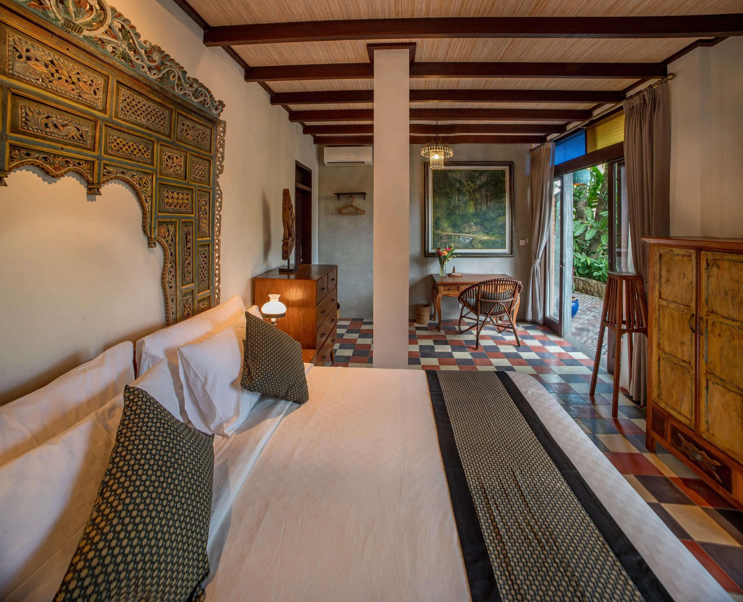 A bedroom with a bed on the left, decorated with pillows, wooden furniture, a painting on the wall, and glass doors opening to a view of greenery outside.