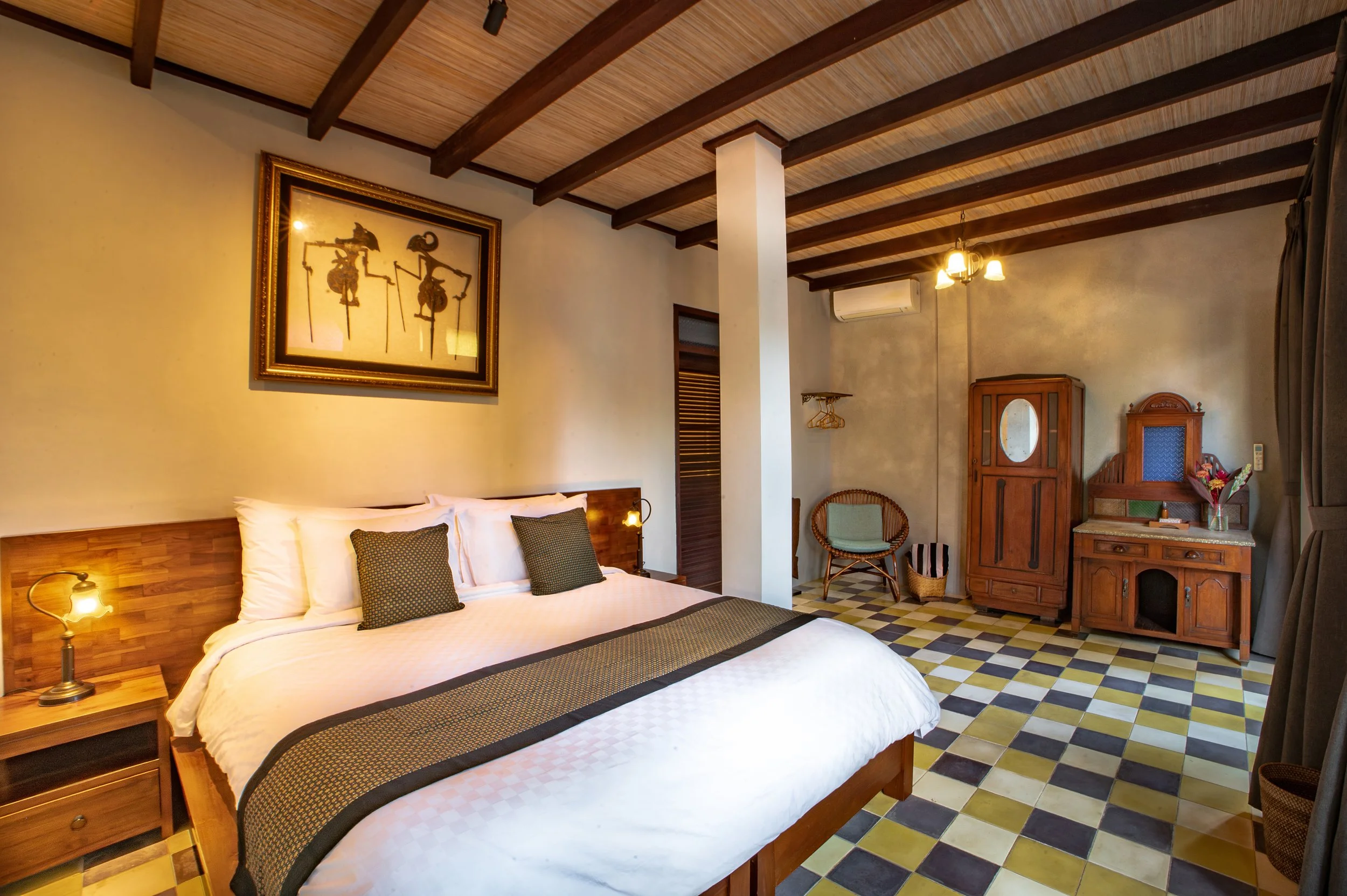 A cozy hotel room with a wooden bed, two pillows, bedside lamp, framed artwork above the bed, wooden furniture, a vintage cabinet, a chair, a wall-mounted air conditioner, and colorful tiled floor.