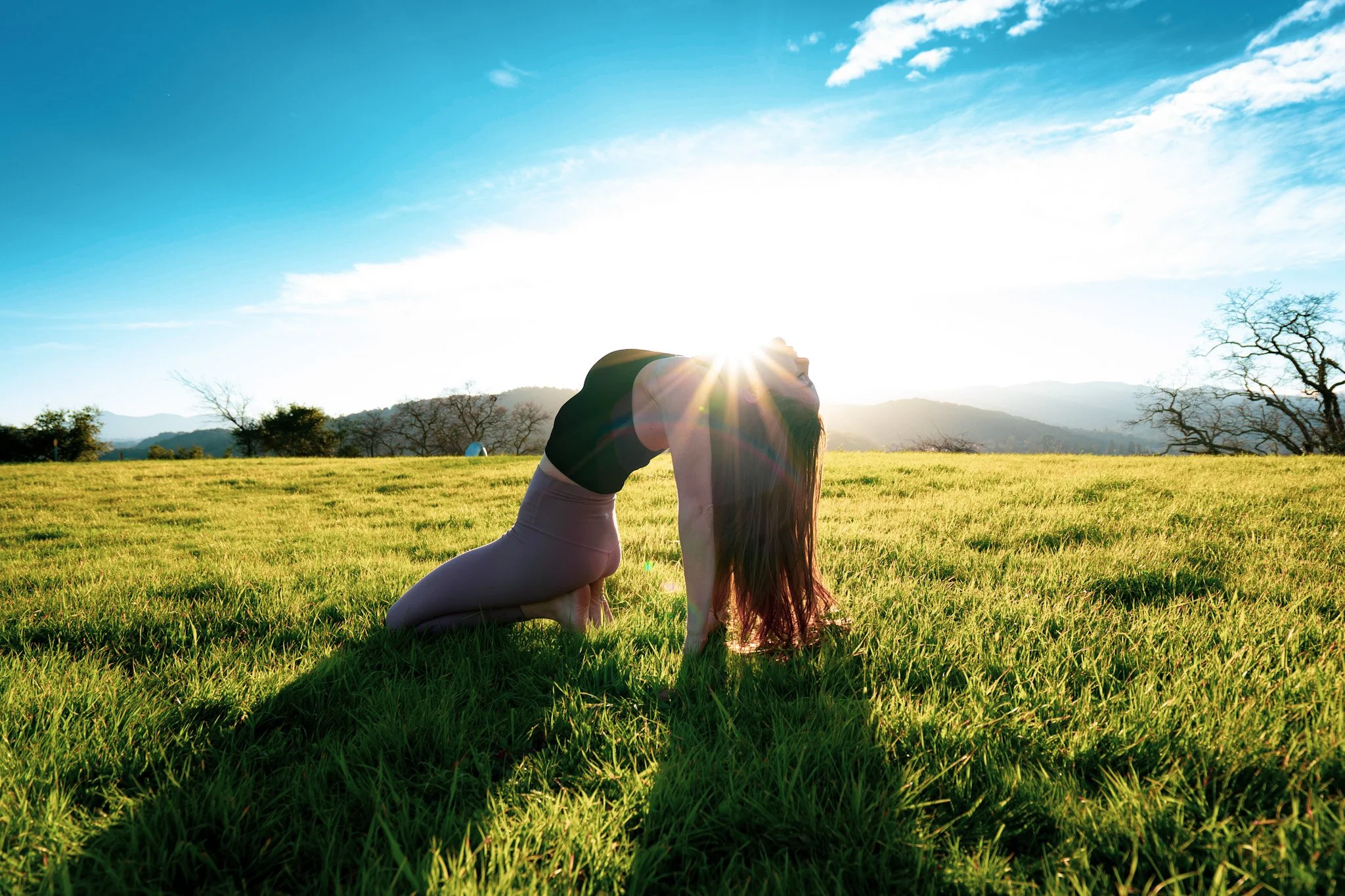 A woman doing yoga outdoors on a grassy field during sunrise or sunset, with hills and trees in the background and a bright blue sky.