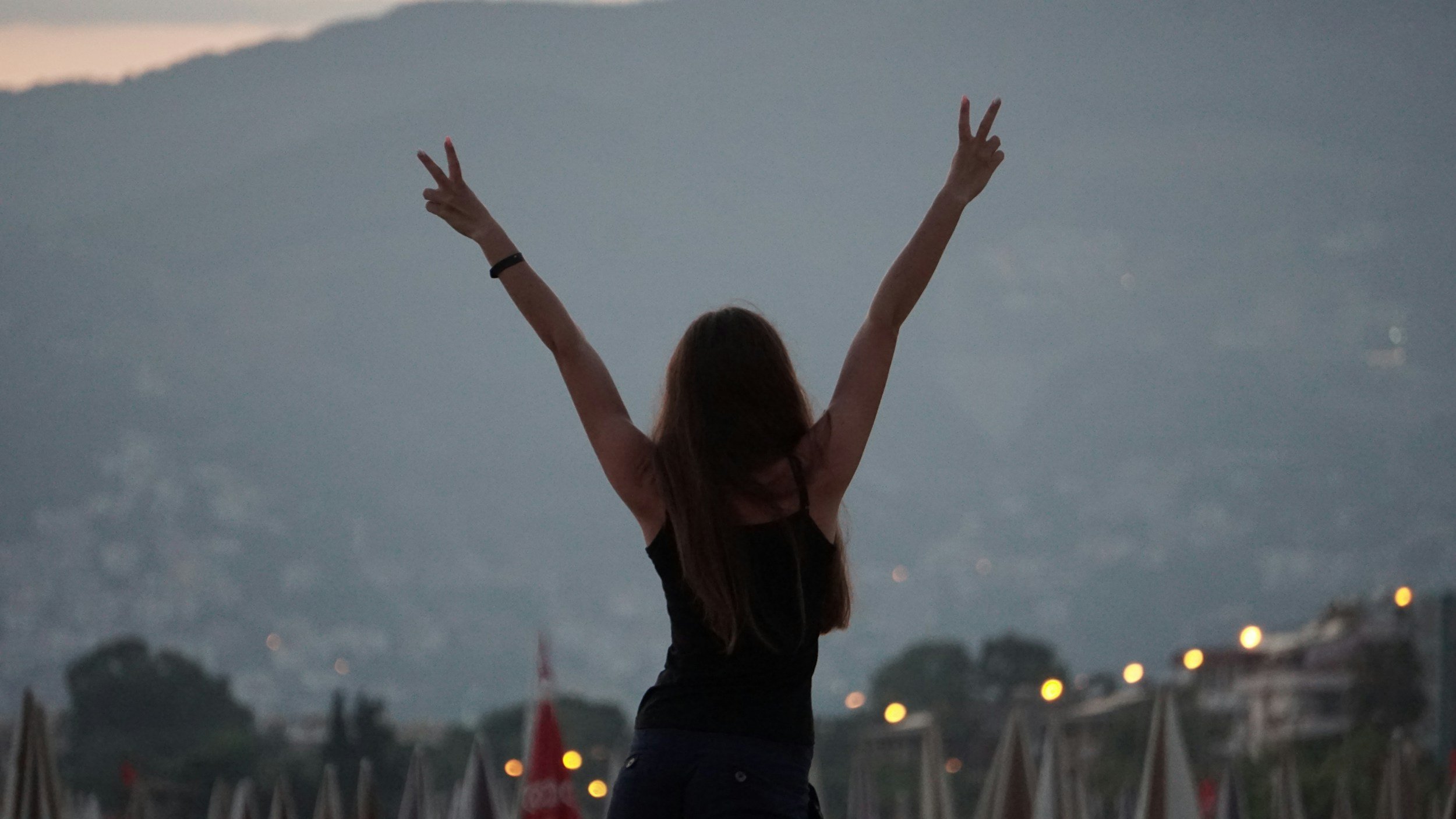A woman with long hair standing outdoors at dusk, raising her arms in a V-shape with peace signs, overlooking a cityscape with blurred lights and mountains in the distance.