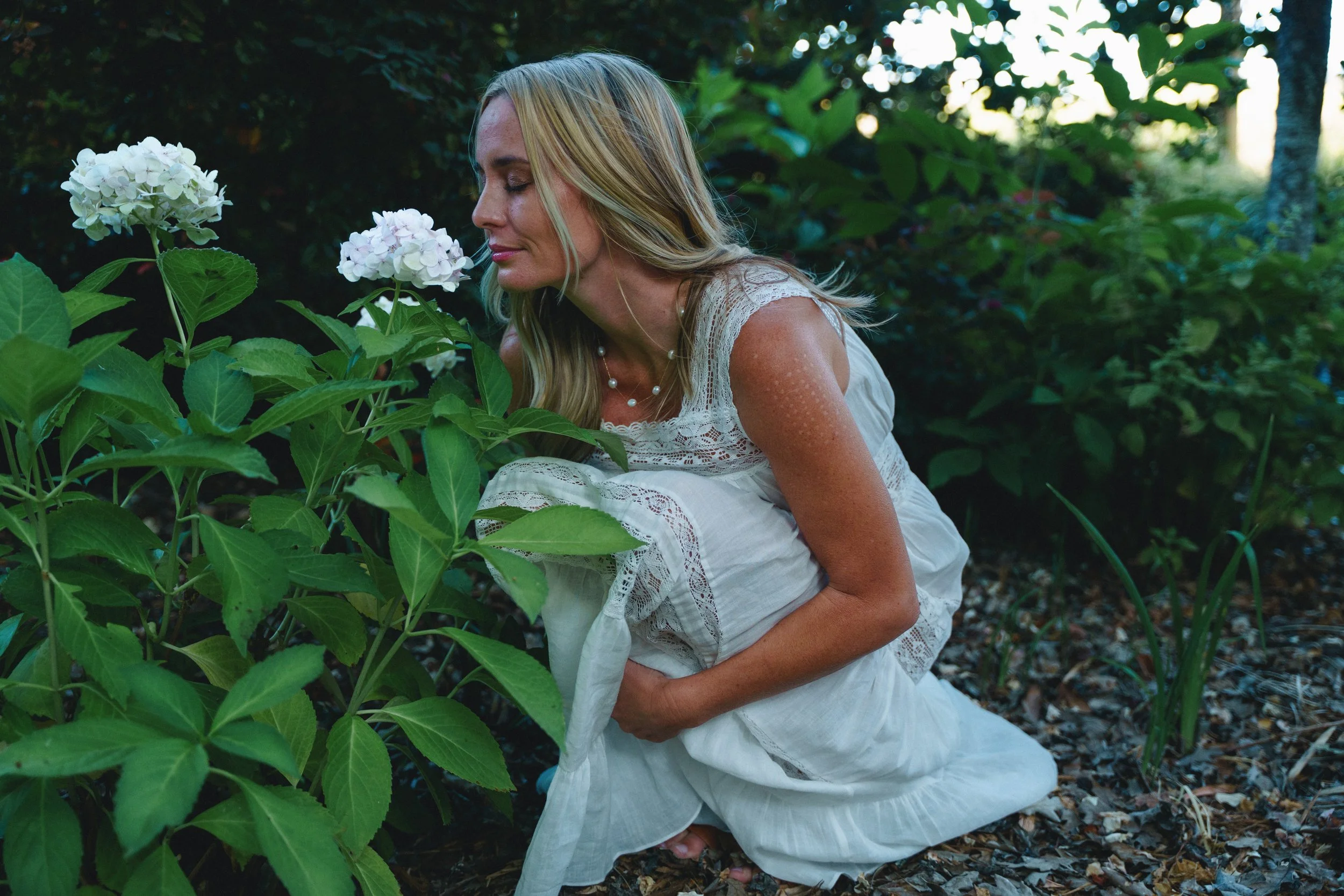 Woman in white dress with lace details sitting among green foliage, holding a hydrangea flower close to her face with eyes closed.