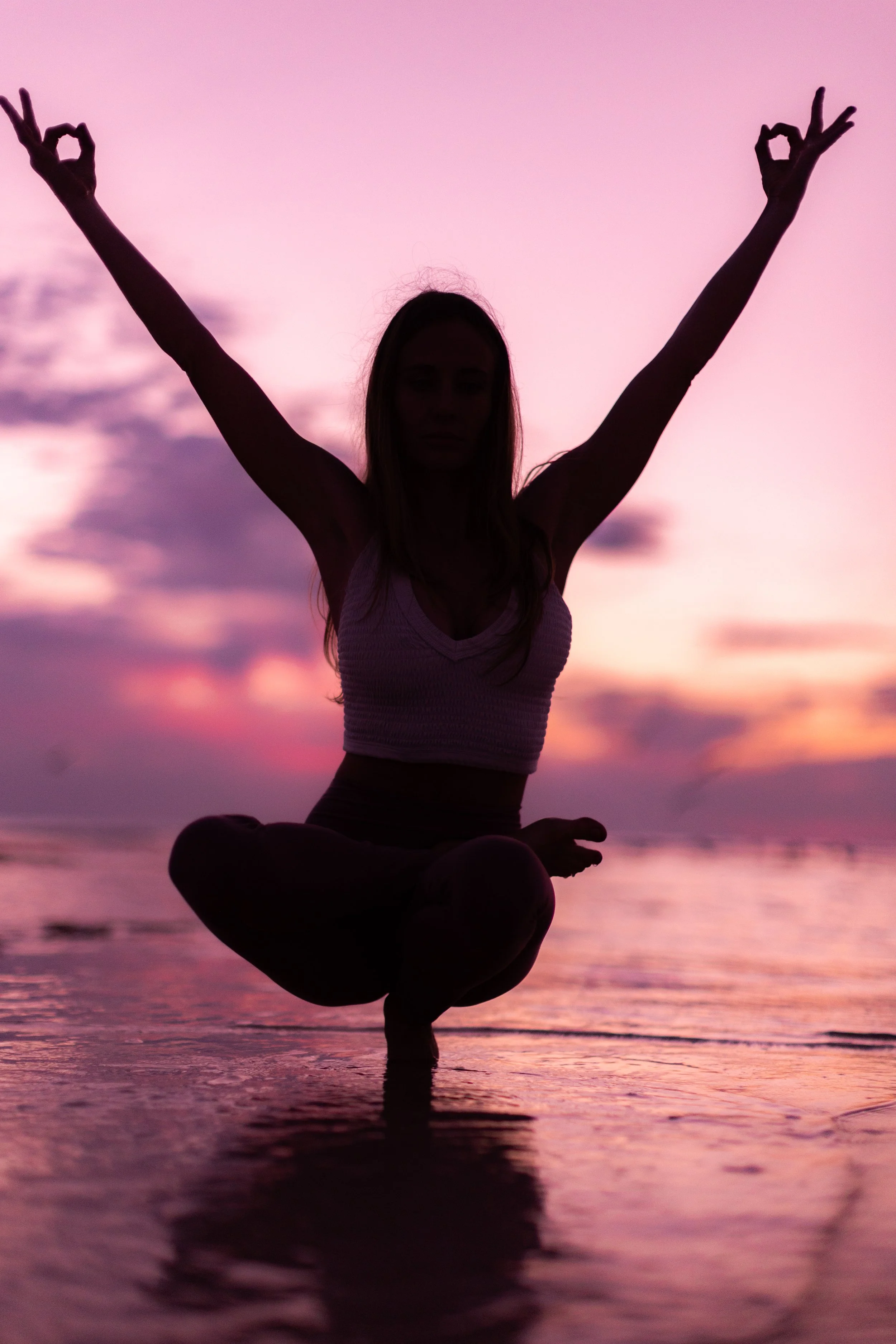 Silhouette of a woman in a yoga pose, balancing on one leg with arms raised and fingers in a mudra, during sunset at the beach with pink and purple sky reflections on the water.