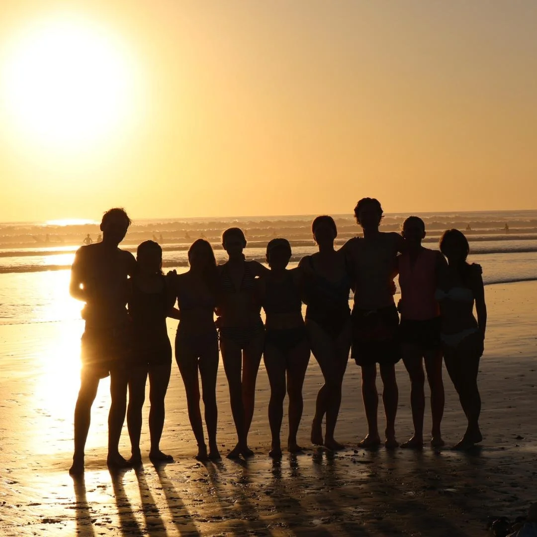 Silhouettes of nine people standing on a beach at sunset, with the ocean in the background.